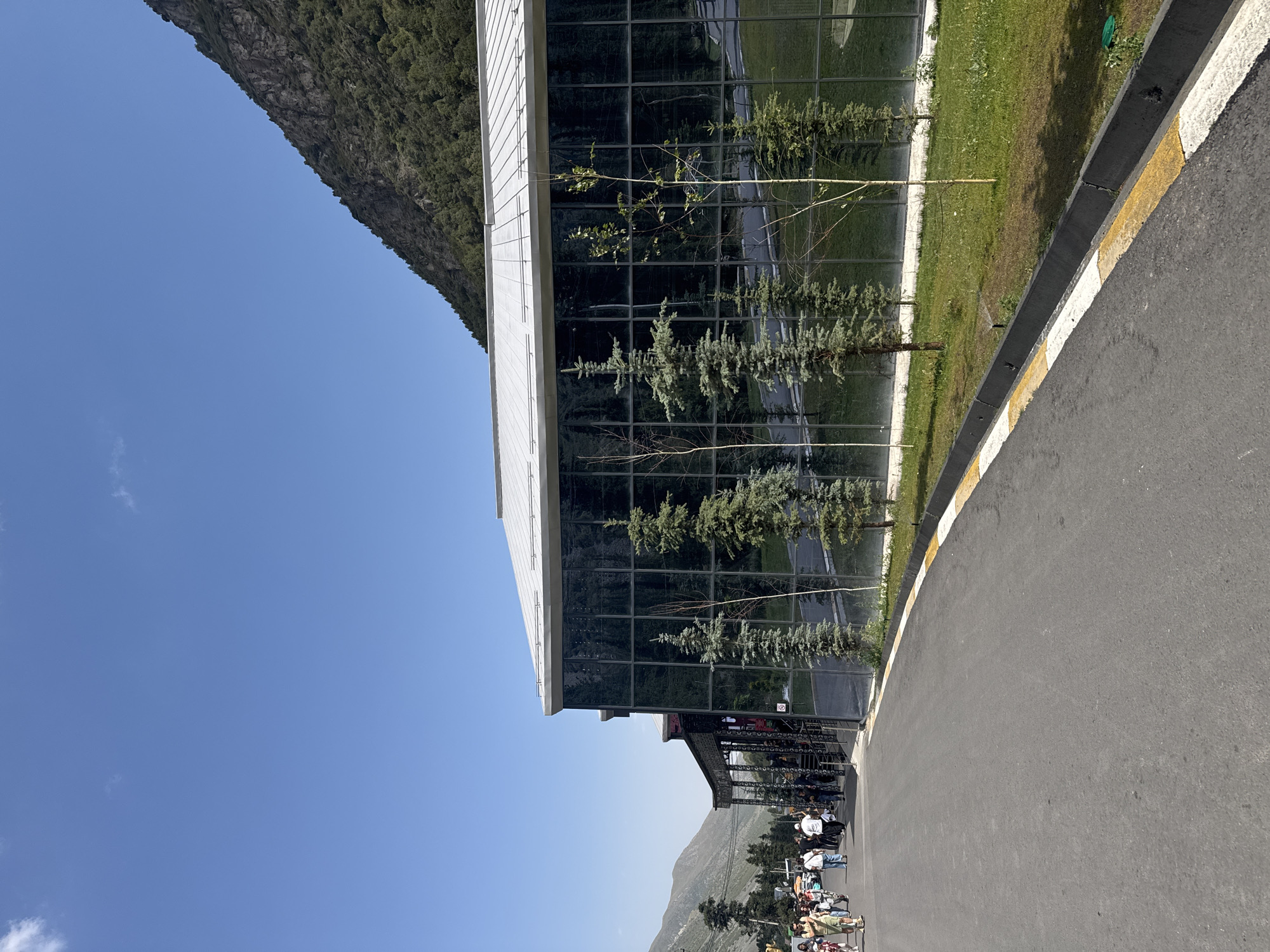 The image captures a wide, paved road in front of a modern building with a large glass facade. The building is situated at the base of a steep, tree-covered mountain under a clear blue sky. A group of people are walking on the road to the left of the building, and several evergreen trees are planted in front of the glass facade. The scene suggests an outdoor area, possibly a tourist destination or a modern facility in a mountainous region.