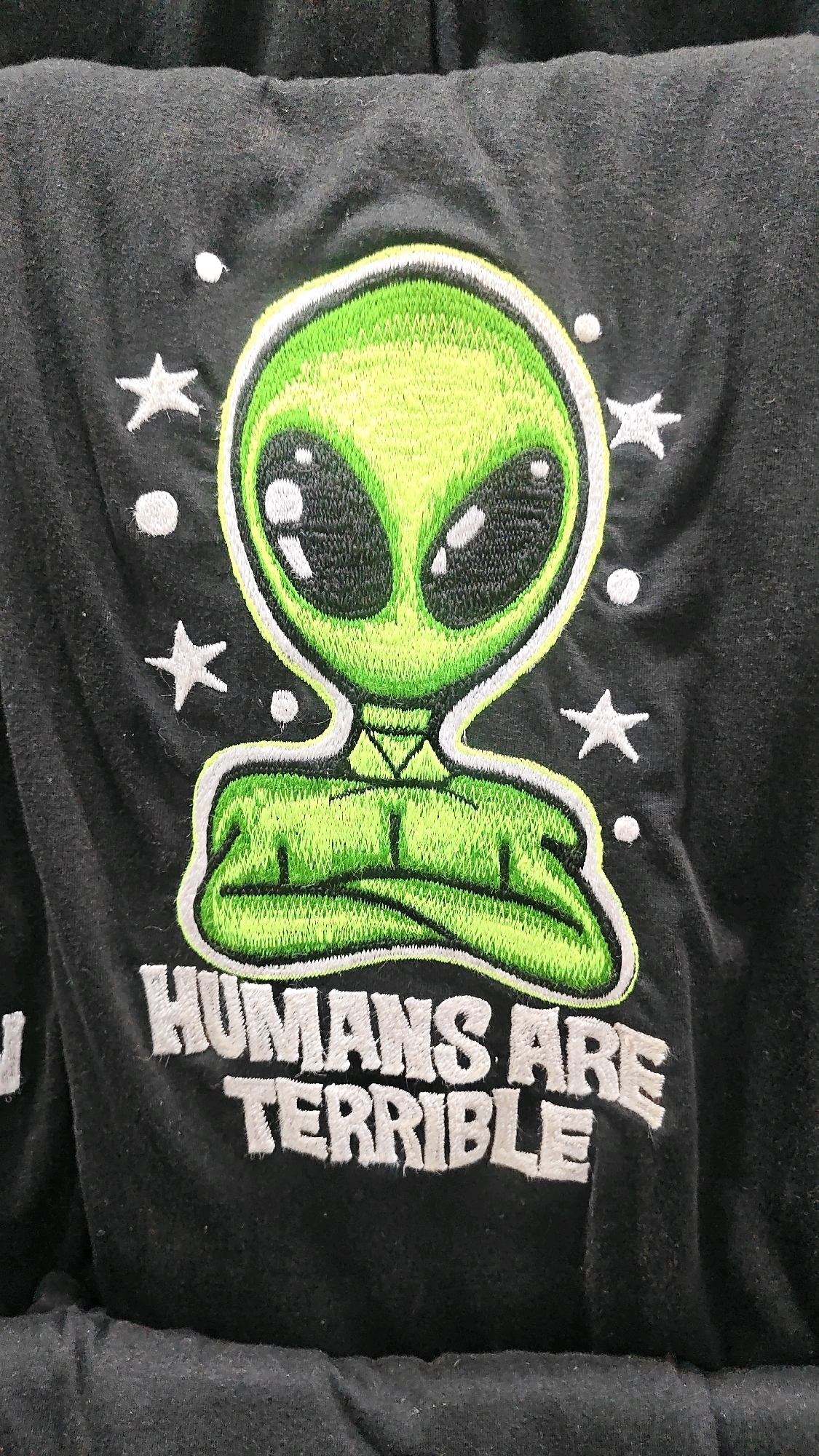 This is a close-up, eye-level, medium shot of an embroidered graphic on a black garment, likely a t-shirt or hoodie. The graphic features a cartoonish, green alien head with large black eyes and a small mouth. The alien's body is depicted as crossed arms. Scattered around the alien are white embroidered stars and dots, resembling a starry sky. Below the alien, white embroidered text reads "HUMANS ARE TERRIBLE". The garment appears to be made of a soft, textured fabric. There is no discernible urban or natural environment in the image, and therefore no indication of its location in Anjuna, India.