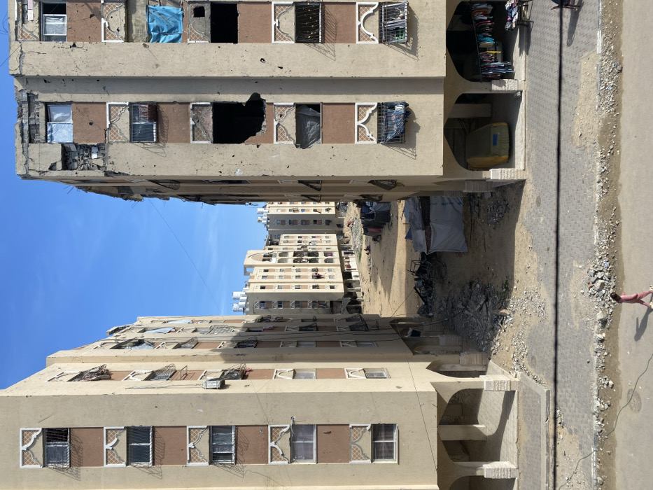 This image depicts a street scene in the city of Khan Yunis, Palestinian Territory, characterized by urban residential architecture under a clear blue sky. Two multi-story buildings, constructed of light tan and brown materials, frame the central view.
The building on the left exhibits multiple windows, many fitted with grates and decorative arch-shaped panels below each window. Some windows show white coverings or hangings. The ground floor features recessed arched openings.
The building on the right is extensively damaged. Its facade is pockmarked with numerous small holes consistent with shrapnel or bullet impacts. A large, irregular black cavity, indicative of an explosion or significant impact, is visible on an upper-middle floor. Other windows are broken, some covered with blue or dark fabric. Laundry hangs from grates on lower windows. The ground floor also has arched openings; one contains a yellow object partially covered, resembling a vehicle, while another displays clothing hanging on a rack.
Between these two foreground buildings, a wide, unpaved dirt and rubble-strewn area extends into the distance. This area contains piles of debris, dark refuse, and several makeshift shelters or tents constructed from tarps and fabric. In the background, more multi-story residential buildings, similar in style but appearing less damaged, line the sides of the street, with water tanks visible on their rooftops.
In the immediate foreground, a paved sidewalk and street are visible. A single individual, dressed in a pink tracksuit, walks from left to right across the street, casting a shadow. Debris is scattered along the edge of the street and sidewalk.