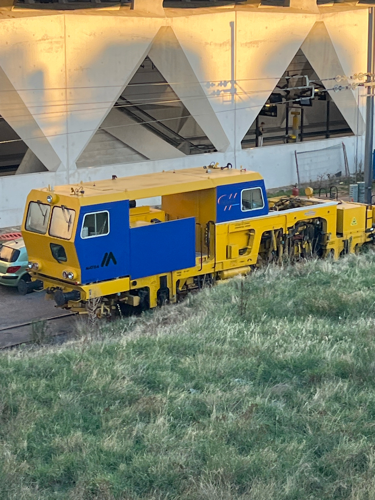 The image shows a bright yellow and blue railway maintenance machine parked on a track next to a grassy slope. The machine is long and appears to be a specialized piece of equipment. In the background, there is a modern concrete structure with large triangular openings, illuminated by a warm, golden light, suggesting either sunrise or sunset. A silver car is parked to the left of the railway machine. The environment appears to be outdoors, with the grass and overhead lighting indicating a potentially urban or industrial setting. The location is identified as Colombier-Saugnieu, France. There are no people clearly visible in the image, but the presence of the car and the railway equipment suggests it is an active or recently used area.