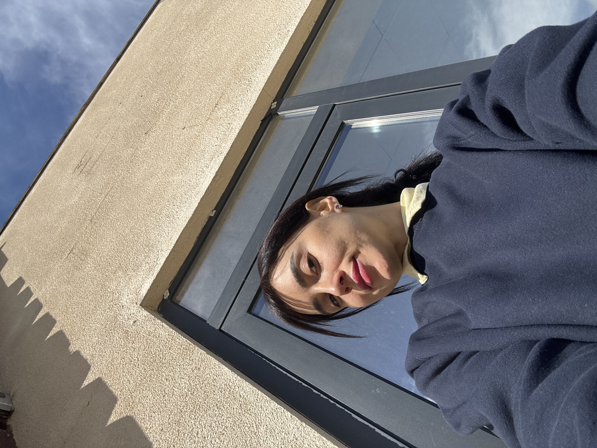 A low-angle shot depicts a person outdoors, likely in the city of Tbilisi, Georgia, as suggested by the architectural style potentially found there. The person is positioned near a building, the corner of which is visible on the left side of the frame. The building has a beige, textured exterior. Next to the building is a window with a dark frame. The person is wearing a dark blue sweatshirt over a light yellow shirt. The sky is visible through the window.