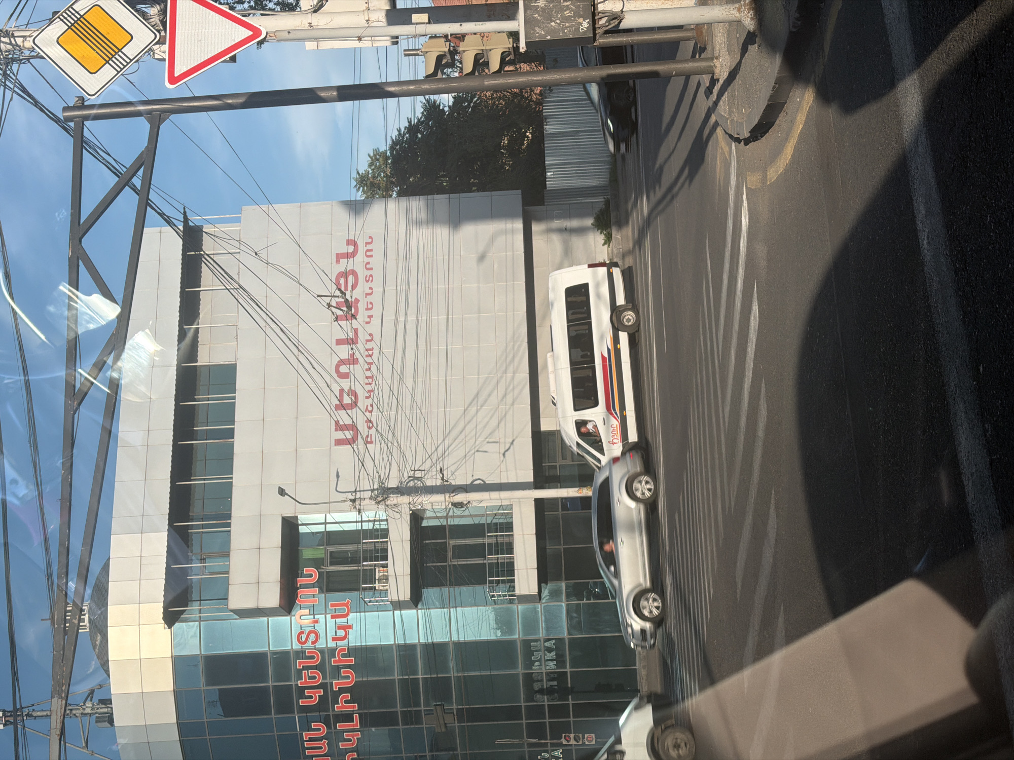 The image captures a street scene in Yerevan, Armenia, on a bright, sunny day. In the foreground, a pedestrian crossing is visible on the asphalt road, with a yellow line bordering the curb. A silver SUV is stopped at the crossing, with its headlights on. To the right of the SUV, a white minivan with orange and blue stripes is also stopped. The minivan has tinted windows and a license plate is partially visible. Behind the vehicles, a tall, modern building dominates the background. The building features a glass facade on the left side, reflecting the blue sky, and a concrete facade on the right. Red Armenian script is visible on both sections of the building. The right side of the building has a gray, grid-like pattern. Above the street, a complex network of electrical wires and poles is strung across the sky. Traffic lights are visible on a pole to the right of the building, with a yellow diamond-shaped sign and a red triangular sign mounted above them. Trees are visible behind the building on the right. The overall scene depicts a typical urban environment in Yerevan, with traffic, architecture, and infrastructure elements.