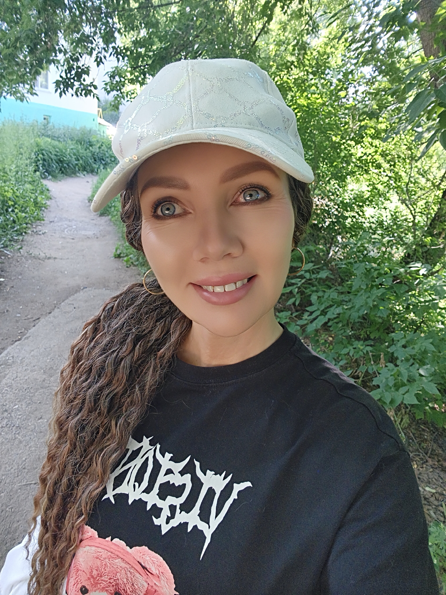 A woman with long, curly brown hair and blue eyes, wearing a white cap and a black t-shirt with a white graphic design, smiles at the camera. She is standing outdoors on a dirt path lined with greenery. To her left, a light blue building is partially visible amongst the trees. The image is taken in a close-up shot, with the woman's face as the primary focus.