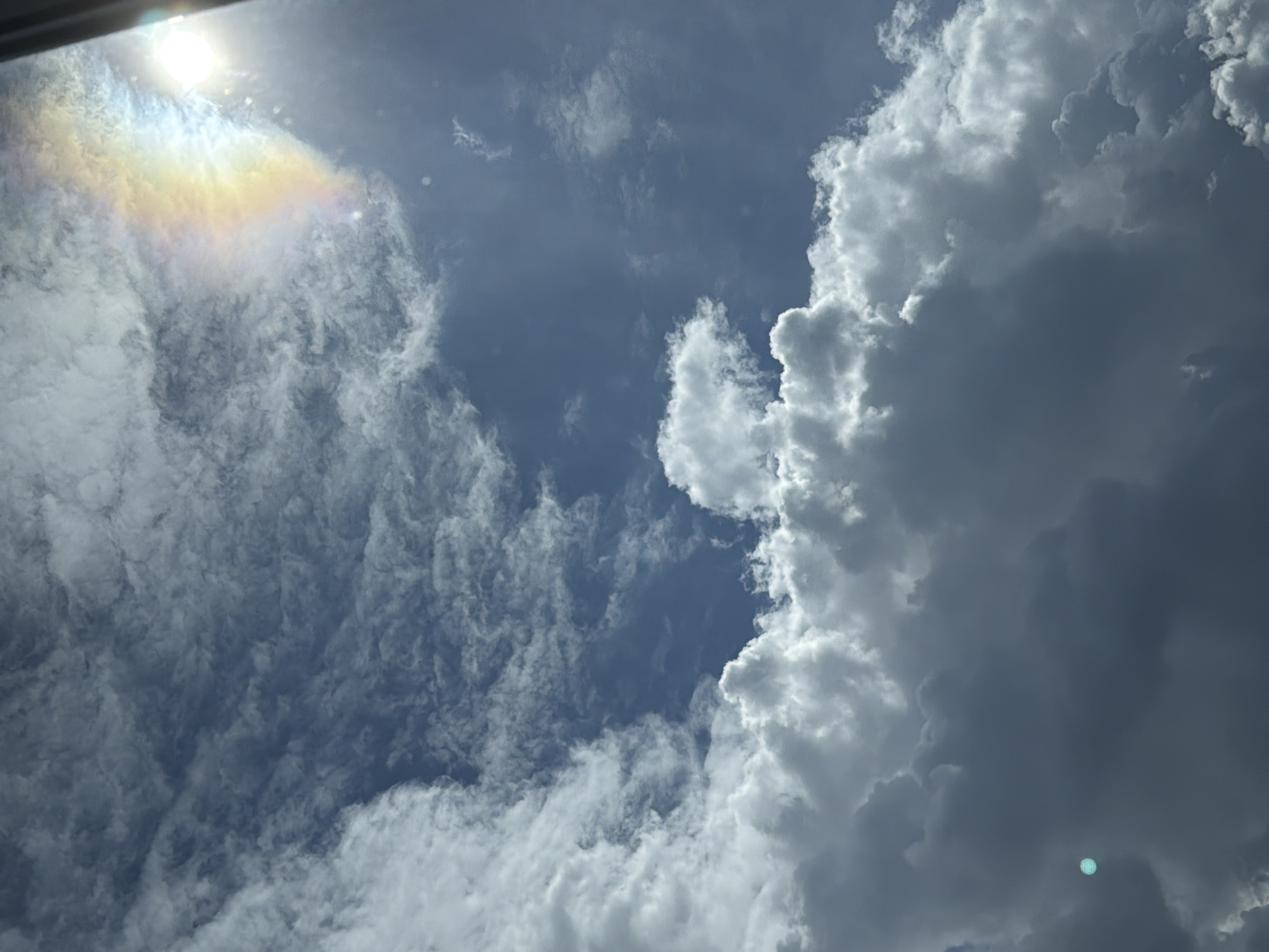 The image captures a dynamic sky scene, predominantly featuring clouds against a blue sky. In the upper right portion of the frame, the sun is visible, partially obscured by clouds, and a rainbow-like optical phenomenon is present, creating a spectrum of colors. The lower part of the image is dominated by large, voluminous cumulus clouds, with some thinner, wispy clouds scattered throughout the upper sky. There is a small, circular, greenish-blue light artifact in the lower left quadrant of the image. The scene appears to be an outdoor shot, looking upwards. There are no people or man-made objects clearly identifiable within the frame, except for what appears to be the edge of a dark structure in the far upper right, possibly a window frame or building edge. The overall impression is a cloudy day with a potential for or recent occurrence of rain, given the presence of cumulus clouds and the sun's rays interacting with atmospheric moisture to create the colorful arc. The location of Perm, Russia, is not discernible from the visual information provided in the image.