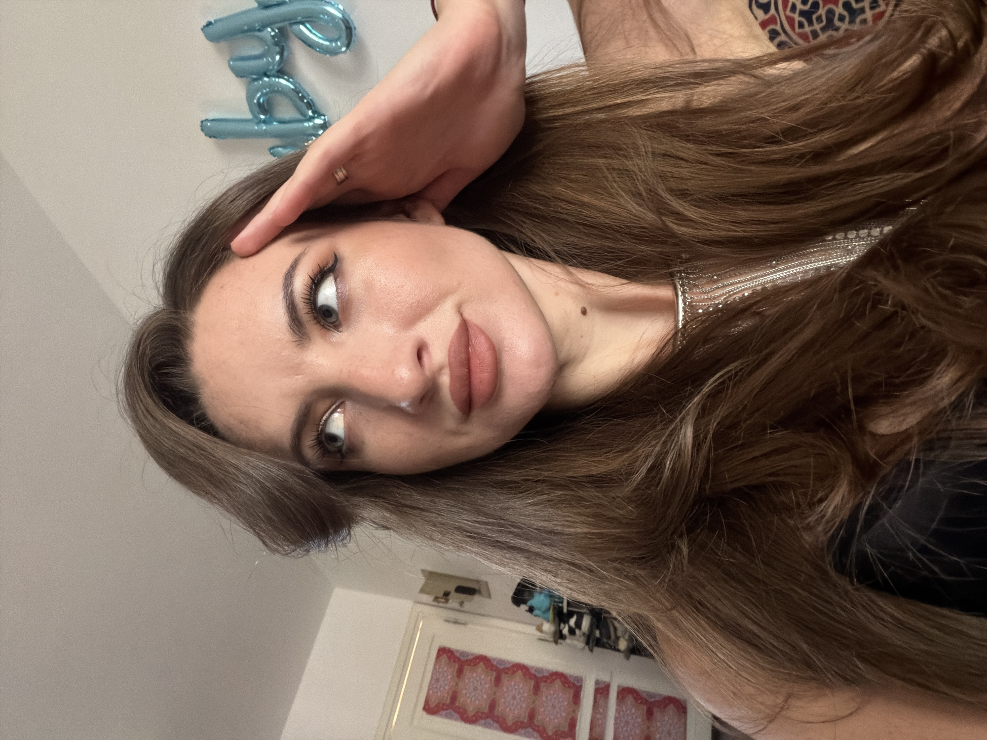 The image shows a close-up portrait of a woman with long, brown hair. She is looking off to the left with her right hand raised to her forehead. Her lips are painted with a matte, nude lipstick, and she has winged eyeliner. A mole is visible on her neck. In the background, a wall with blue balloons spelling out "baby" can be seen. To the left, there is a door with a patterned design. The scene appears to be indoors, likely in a residential setting. The lighting suggests natural light coming from the left. The location of Sharm El Sheikh, Egypt, is not evident in this image.