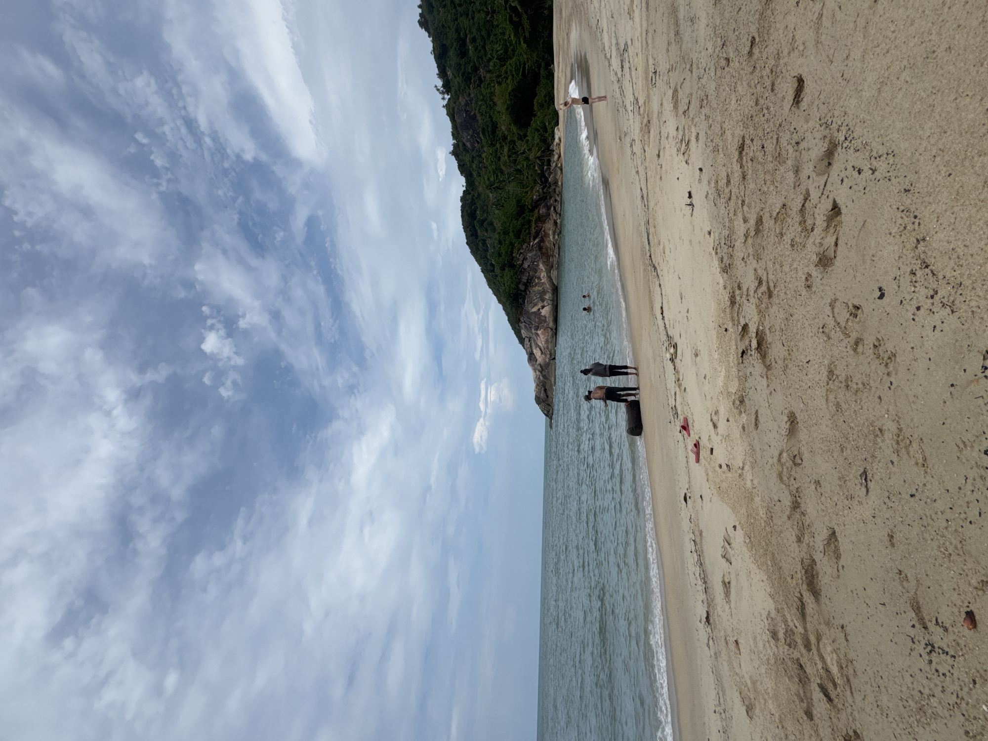 The image captures a beach scene in Baan Thong Nang, Thailand. The view is oriented with the sea on the right and the sky to the left. A sandy beach occupies a significant portion of the foreground, displaying footprints and scattered debris, including a pair of red flip-flops.
A small group of people is visible along the shoreline, some standing near the water's edge. A rocky headland densely covered with green vegetation extends into the sea. The sky is characterized by a mix of white and grey clouds. The sea appears calm.