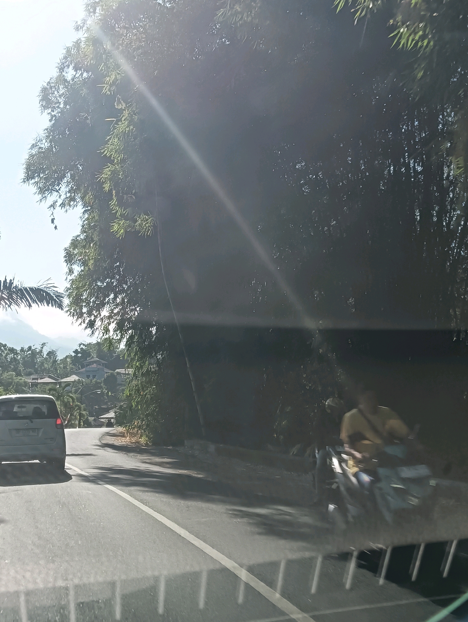 The image depicts a road scene in Tomohon, Indonesia, characterized by a bright, sunny day with sun glare. On the left side of the road, a white car is visible from the rear. The road itself is paved with white lane markings. To the right of the road, a dense cluster of bamboo trees creates a shaded area. Further down the road, a person on a motorcycle, wearing a yellow shirt, is moving away from the viewer. In the background, residential buildings are scattered on a hillside, with lush green vegetation and distant mountains under a clear blue sky.