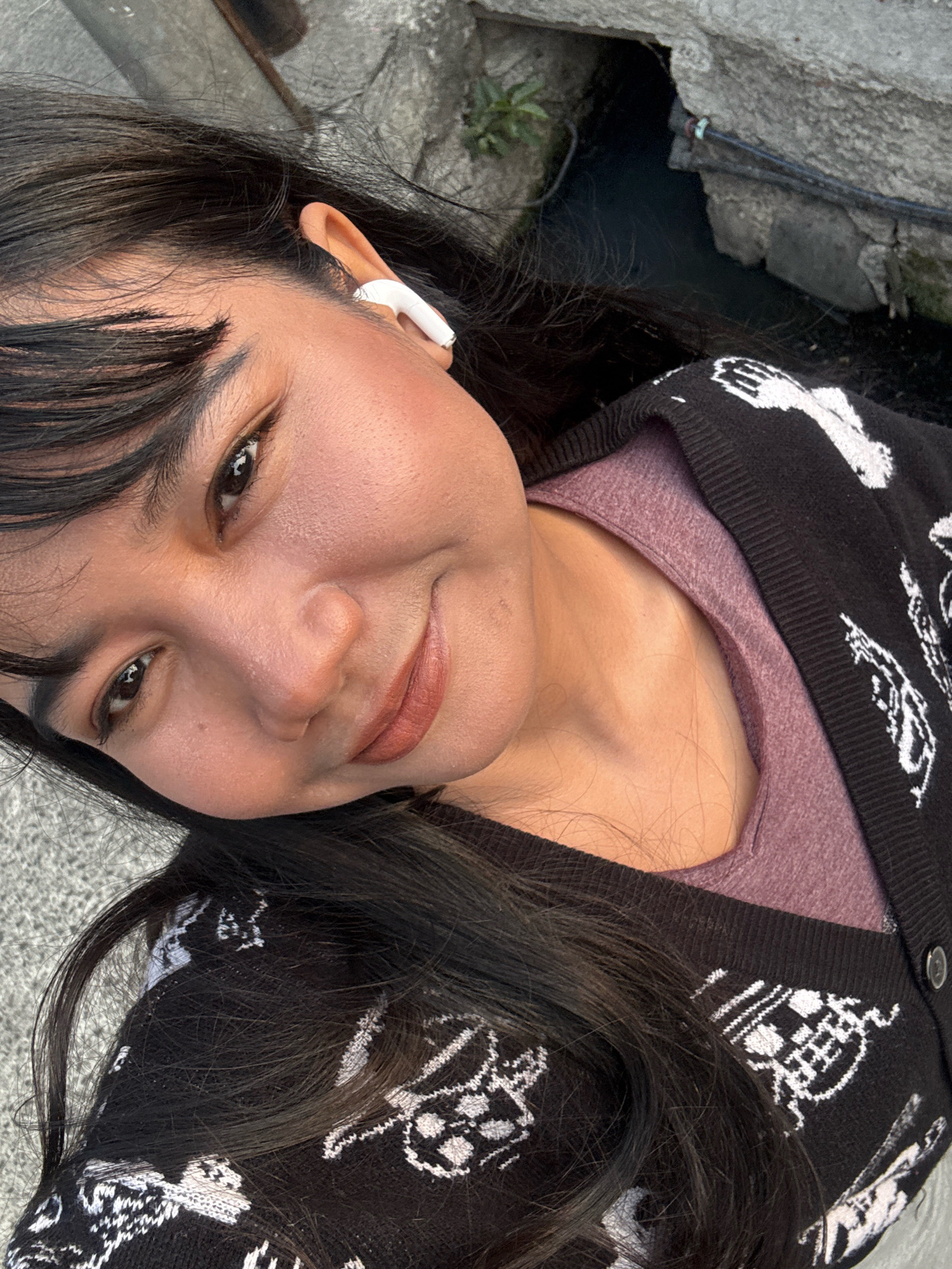 A medium-close-up, overhead selfie shows a woman with dark hair and bangs wearing a purple shirt and a black cardigan with a white skull pattern. She is smiling and looking at the camera, with a white earbud in her right ear. The background shows a stone wall and a dark, narrow channel of water. The scene is outdoors, and appears to be in an urban environment.