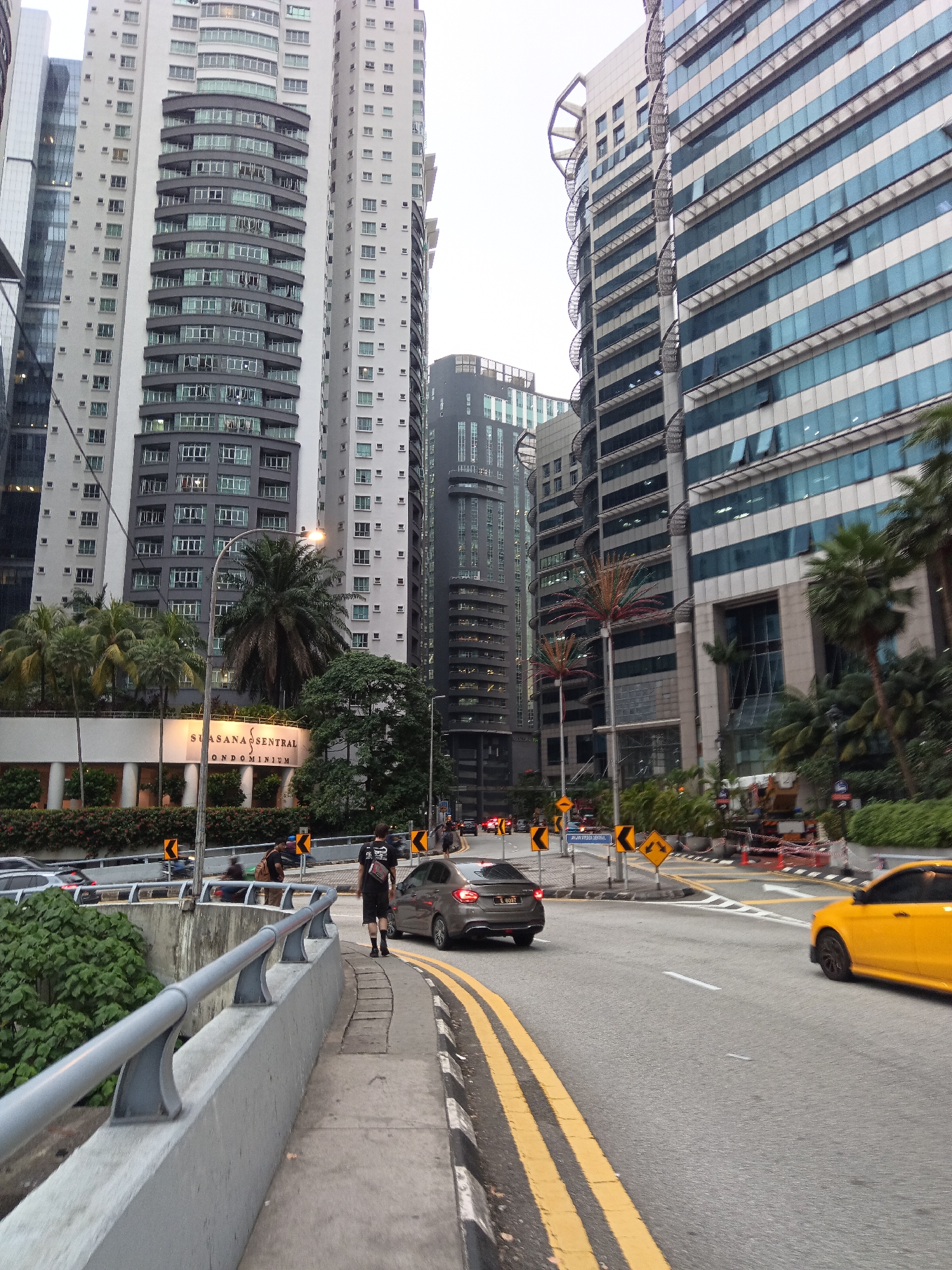This is an eye-level, medium shot of a street scene in Kuala Lumpur, Malaysia. The perspective is looking down a curved road lined with modern skyscrapers. On the left side of the frame, there are several tall residential buildings with white facades and dark, curved balconies. In the center, a black car is driving away from the camera, and a man in a black t-shirt and shorts is walking on the sidewalk in the foreground. A yellow car is visible on the right side of the road, also driving away. The road is marked with double yellow lines and white lane dividers. Traffic signs indicating curves and lane changes are present. In the background, there are more contemporary office buildings with blue-tinted windows. The sky is overcast, suggesting it may be daytime or late afternoon. Lush green foliage and palm trees are visible near the base of the buildings on the left.