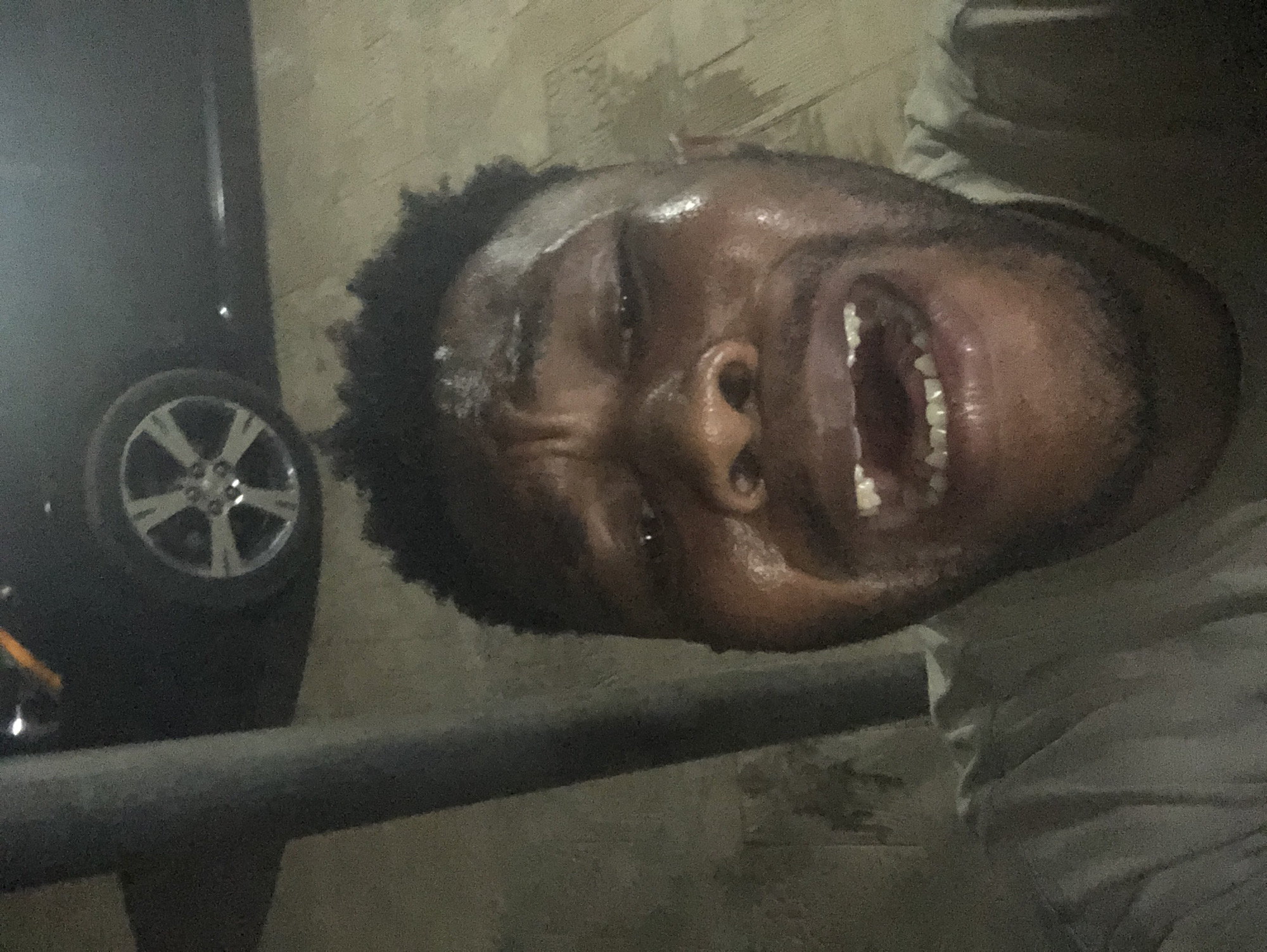 The image appears to depict a man with dark skin and short, curly hair, with a sheen of moisture on his forehead. His mouth is open, revealing his teeth. He is wearing what seems to be a light-colored shirt. Adjacent to him on the left side of the frame is the tire of a car, with a silver rim. A metal bar is visible in the foreground, and a textured wall is in the background. The setting is an urban area with a car. The location is Unknown, Unknown.
