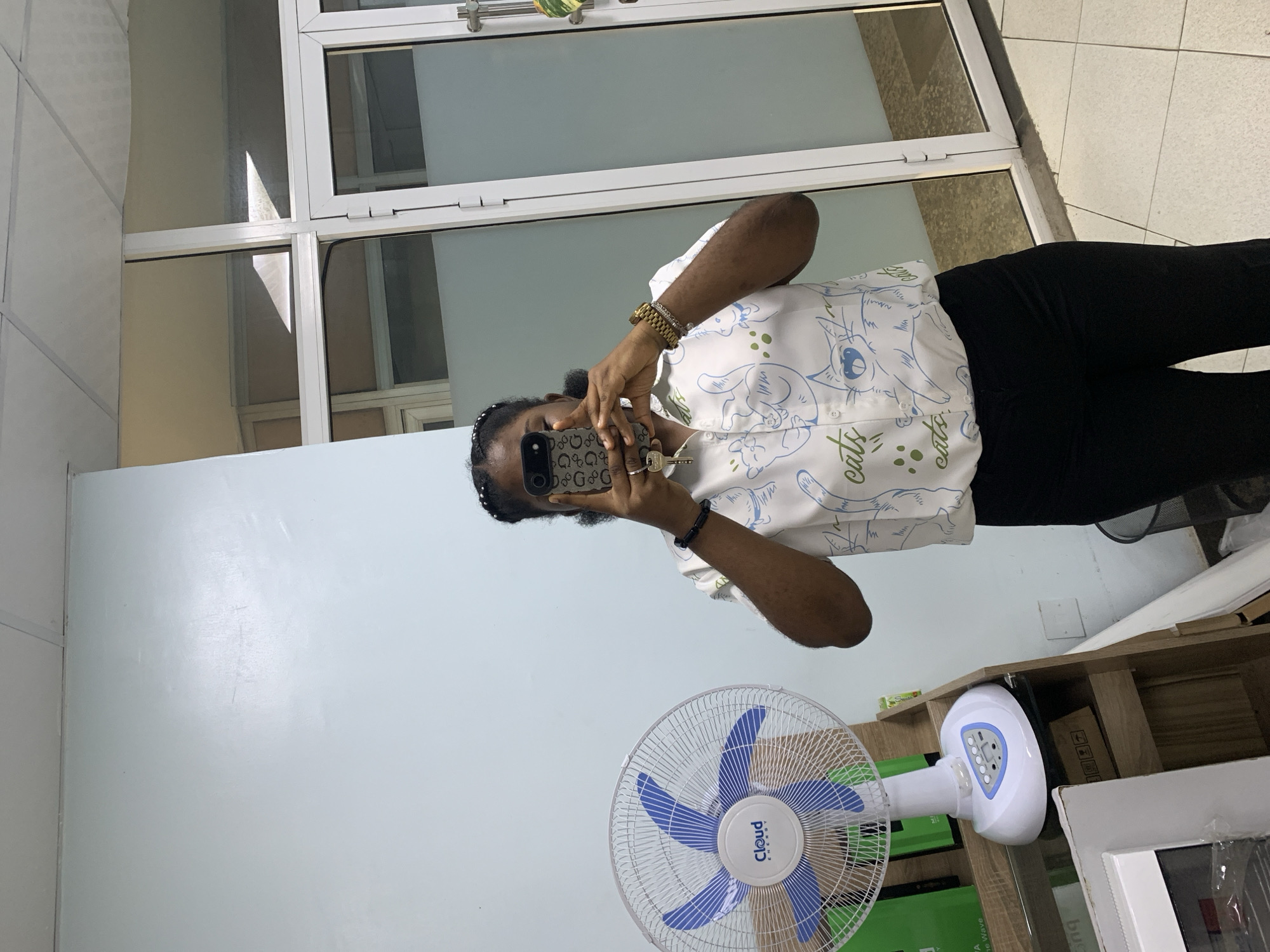 A young Black woman is standing in an indoor space, possibly an office or shop, in Ikeja, Nigeria, taking a mirror selfie. She is wearing a white button-up shirt with a pattern of blue and green cartoon cats and the word "cats" repeated, and black pants. She has dark hair styled in braids with some strands loose around her face. She is holding a smartphone in her right hand, using it to capture her reflection. A large white and blue oscillating fan is positioned to her left on a wooden shelf unit. Behind her, to the right, is a glass-paned door and wall, with reflections visible in the glass. The wall to her left is a light blue. The floor is tiled.