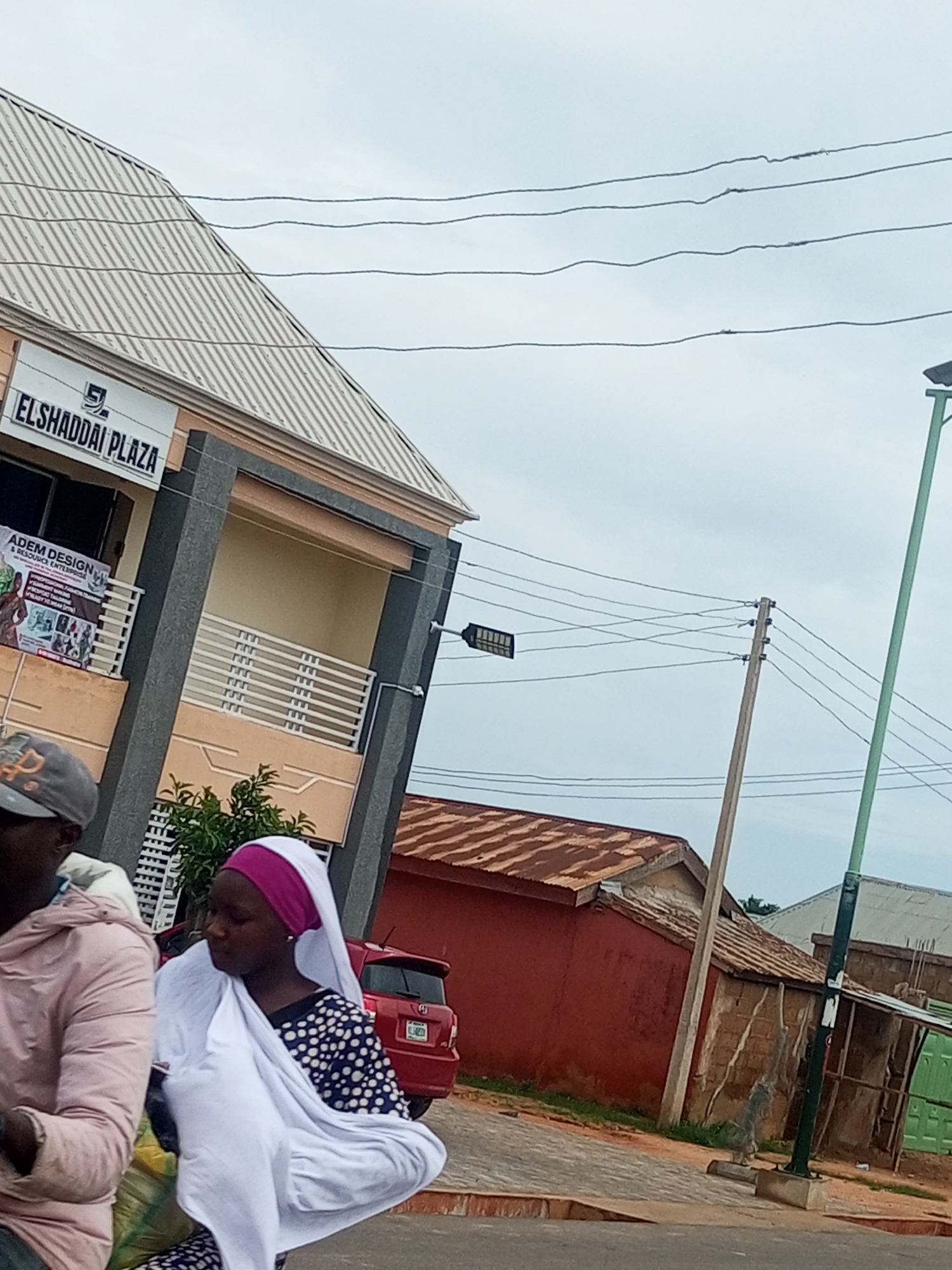 The image captures a street scene in Tempure, Nigeria. In the foreground, two individuals are partially visible, one wearing a pink hooded jacket and a cap, and the other a woman in a dark polka dot dress with a magenta and white headscarf. She is draped in a white cloth, possibly a hijab. Behind them, a multi-story building with a corrugated metal roof is visible, featuring signage that reads "ELSHADDAI PLAZA." Below this sign, a banner advertises "ADEM DESIGN & RESOURCE ENTERPRISE" with text describing their services.
Further back, a red car is parked, and beyond that, a red building with a rusty corrugated metal roof stands adjacent to other structures. Utility poles with overhead wires crisscross the sky, and a green lamppost is also present. The ground appears to be paved with interlocking stones. The overall atmosphere suggests an urban or semi-urban setting under an overcast sky.