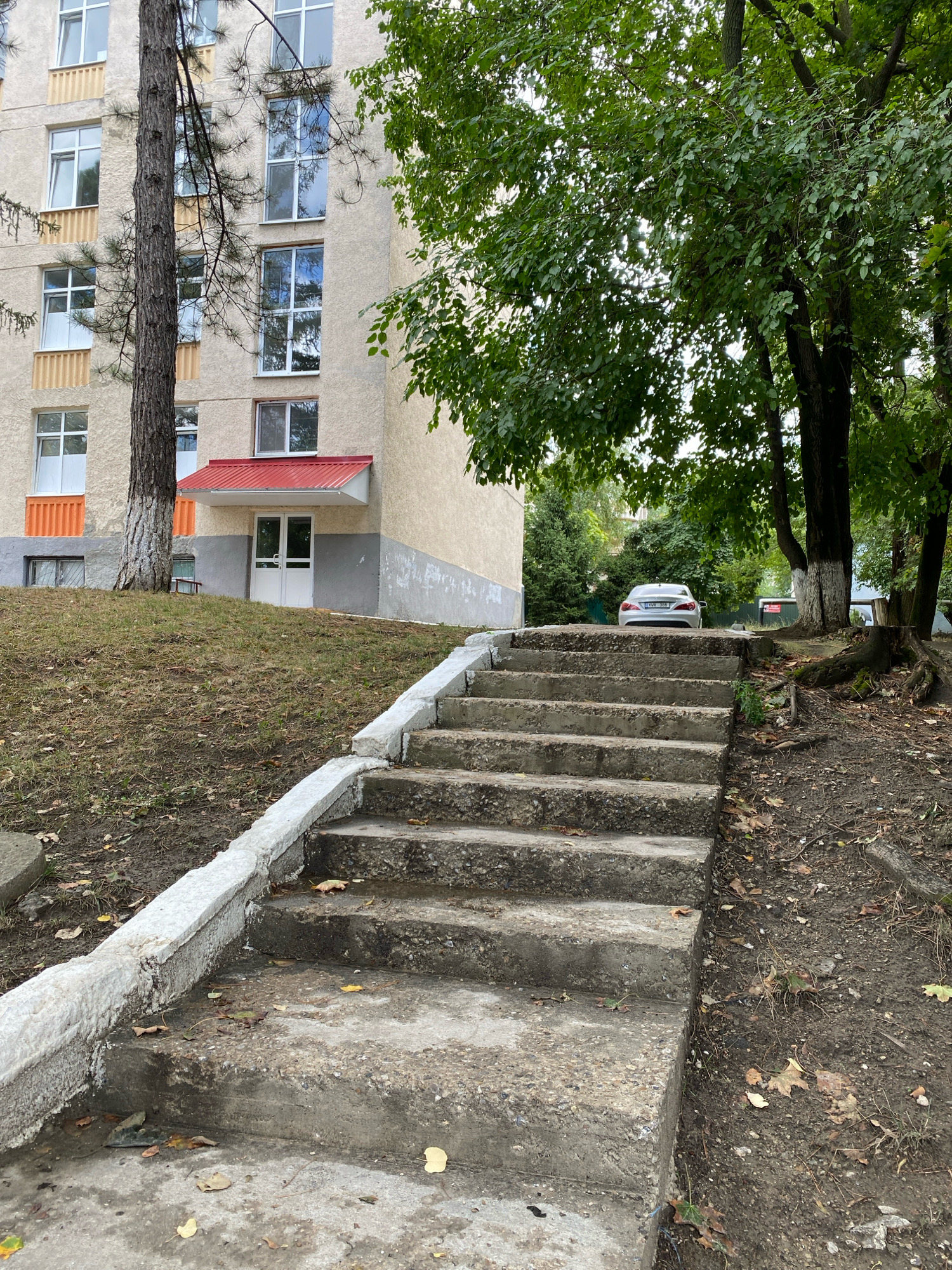 The image depicts an outdoor scene in Chișinău, Moldova. In the foreground, a set of concrete stairs with a white railing leads upwards. The stairs are worn and some fallen leaves are scattered on them. To the left of the stairs, a grassy slope with some dry patches and scattered leaves is visible, leading towards a multi-story building. The building has a beige facade, numerous windows with white frames, and some orange-colored sections. A red awning covers a double white door at the base of the building. To the right of the stairs and behind the building, a white car is parked on a paved area, partially obscured by trees with lush green foliage. The environment suggests a residential area or an institutional setting with mature trees and vegetation.
