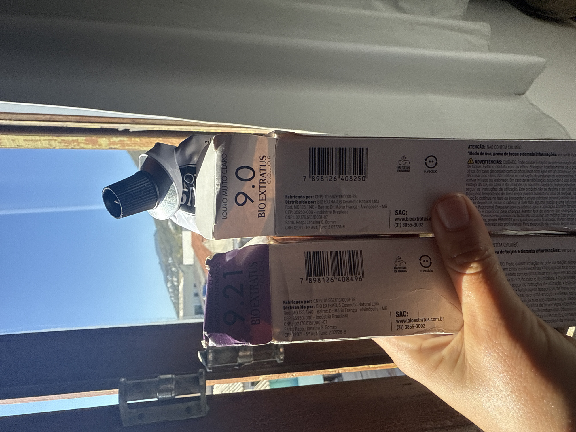 The image shows two boxes of hair dye held in a person's hand in front of a window. The boxes are labeled "9.21 BIOEXTRATUS COLOR" and "9.0 BIOEXTRATUS COLOR". The window looks out onto a sunny, blue sky with some distant hills and buildings, suggesting an outdoor or semi-outdoor setting in a town. The overall impression is of a close-up shot, with the focus on the hair dye boxes.