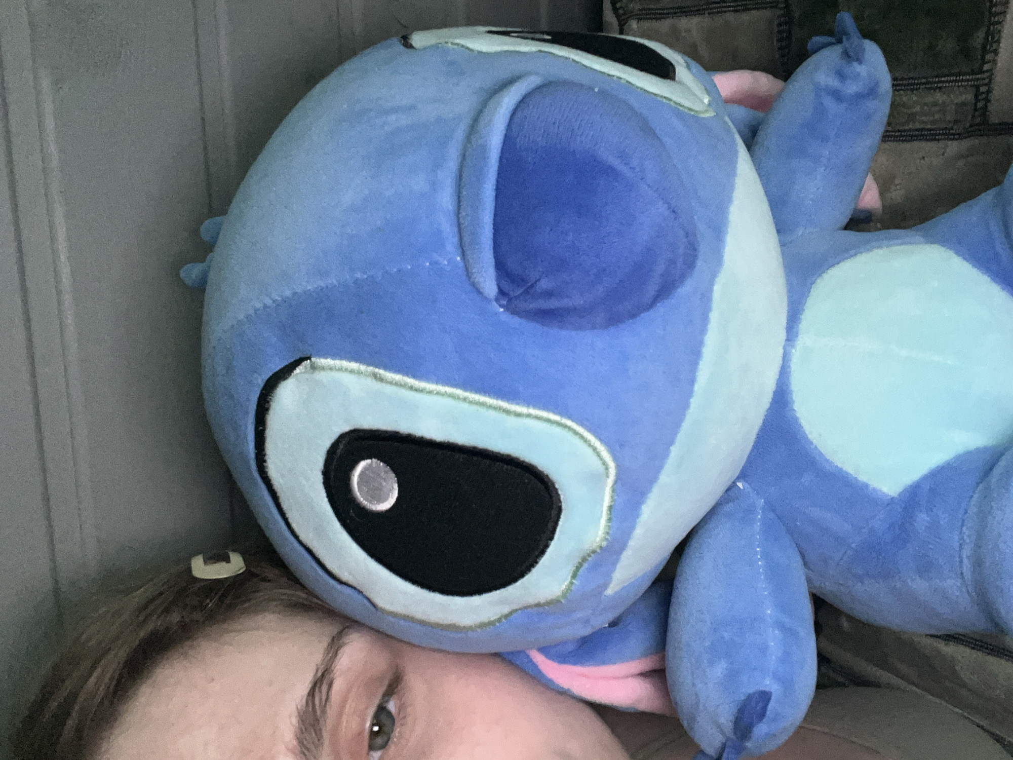 A close-up, top-down, selfie-style photograph shows a person's left eye and forehead, partially obscured by a blue Stitch plush toy. The eye is green with a light brown iris and appears to be partially closed. A light-colored hair clip is visible in the person's brown hair near the temple. The Stitch toy is large and plush, with a light blue body, darker blue accents, and a light blue stomach patch. One of Stitch's large black eyes is prominent, with a silver reflective pupil. The other eye is partially closed, showing a blue fabric cover. Pink fabric accents are visible on Stitch's ears. The background appears to be a muted, textured surface, possibly a couch or wall. The scene is indoors, with soft lighting. The location of Ashkadar, Russia is not depicted or suggested in this image.