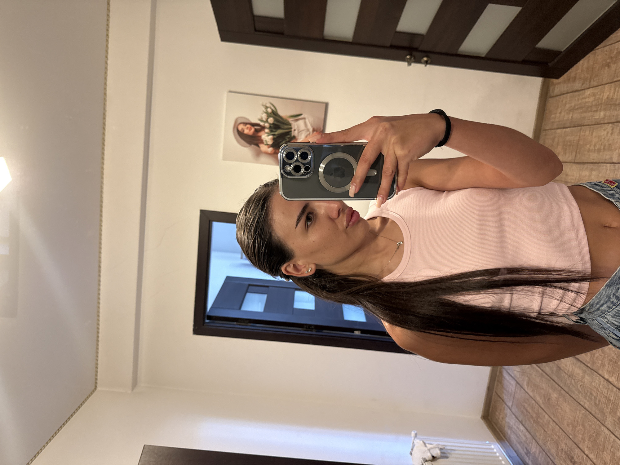 The image shows a young woman with long dark hair pulled into a ponytail, taking a mirror selfie indoors. She is wearing a pink ribbed tank top and denim shorts. Her right arm is raised, holding a dark-colored smartphone with a clear case, displaying a prominent Apple logo. On the wall behind her, to her left, is a framed picture of another young woman wearing a white hat and holding a bouquet of tulips. To her right, a dark wooden door with frosted glass panels is visible. The room features light-colored walls, a decorative border near the ceiling, and a wooden floor. A white radiator is partially visible on the left wall. The overall impression is that of a domestic setting, possibly an apartment or house. There is no information in the image to indicate a specific location within the city of Chișinău, Moldova.