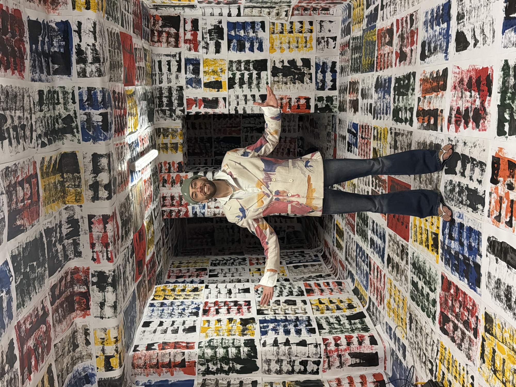 This image depicts a man posing within a distinctive art installation in Toulon, France. The man, appearing to be in his late 20s or early 30s with a beard and a green beanie, is dressed in a white t-shirt, a light-colored jacket adorned with abstract paint splatters, blue jeans, and white flip-flops. He stands with his arms slightly outstretched, smiling and looking directly at the camera, conveying a sense of enjoyment and engagement with his surroundings.
The entire enclosed space – walls, ceiling, and floor – is covered with numerous rectangular sheets of white paper. Each sheet features an abstract print characterized by textured, block-like applications of paint in black, red, yellow, and blue. These prints create a dense, colorful, and repetitive pattern throughout the room, giving the impression of an immersive art experience. A fluorescent light fixture is visible on the left side of the room.
The photograph is rotated 90 degrees clockwise, ingeniously making it appear as though the man is standing upright on a side wall, looking into a seemingly deep and narrow corridor. This clever manipulation of perspective transforms the ordinary room into an illusion, playing with the viewer's sense of gravity and space, indicative of an interactive art exhibit. No discernible text is visible within the image.