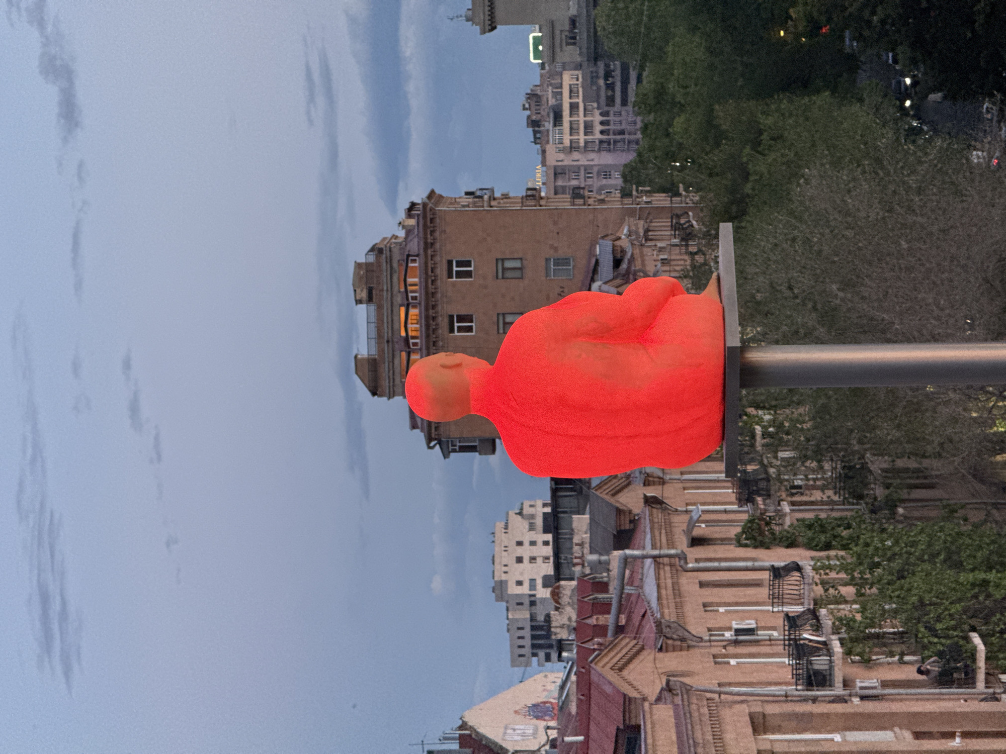 An eye-level, vertically oriented, daytime shot shows a red sculpture in the foreground with a city scene as background. In the foreground stands a large, solid red statue of a seated figure. The figure has no distinct facial features, but is seated with its legs crossed and arms resting on its knees. It sits atop a gray metal pole, appearing to be a modern art installation.
The background features a cityscape. A mix of old and newer buildings are visible, mostly constructed from brick and concrete. Some buildings have traditional architecture with balconies, while others are more modern with clean lines and large windows. There are scattered trees, filling the spaces between the buildings and adding some greenery to the urban landscape. The sky is slightly overcast, with light clouds spread evenly across the frame.
The location is the city of Yerevan, Armenia.