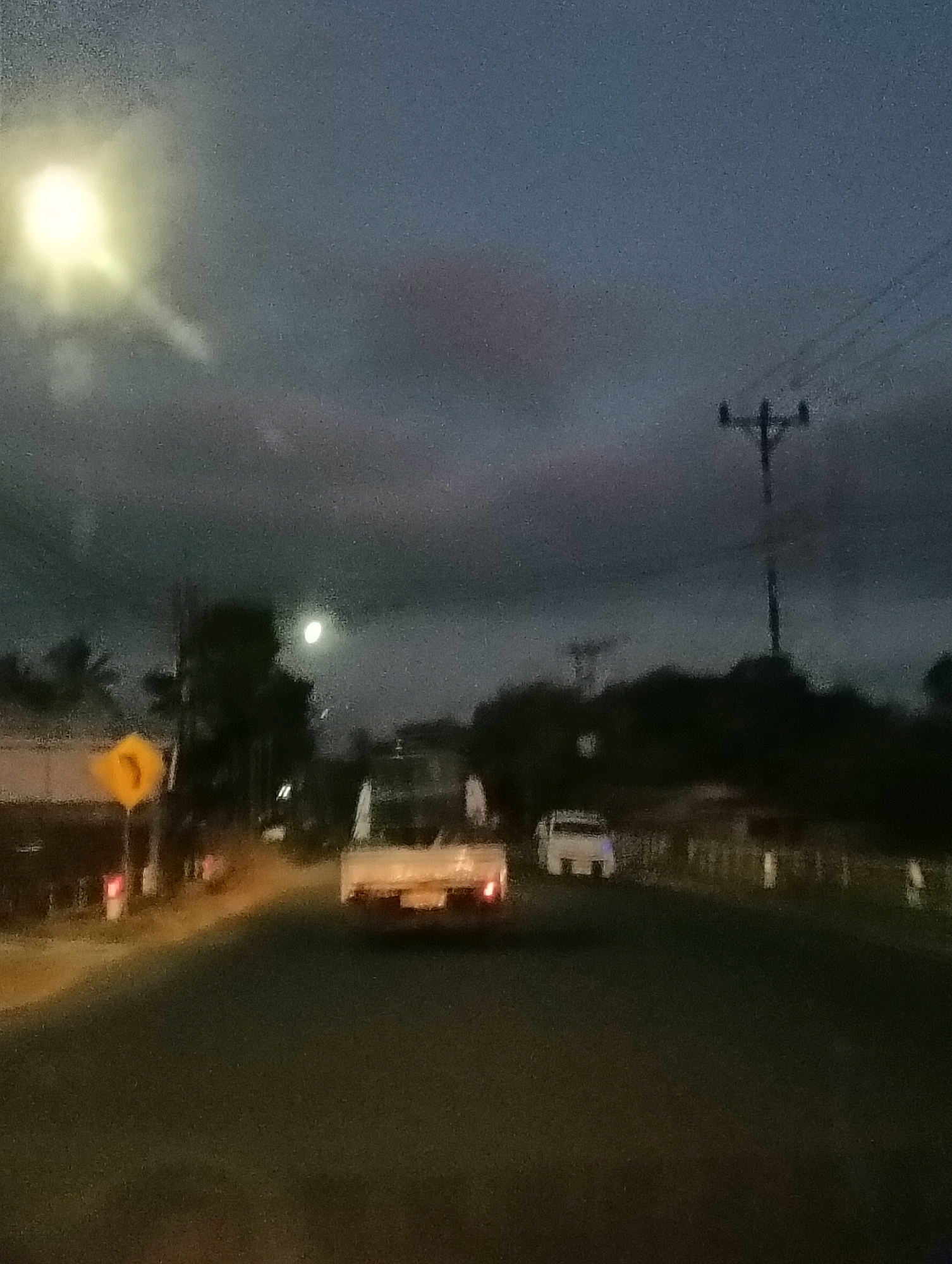 A nighttime view from a vehicle shows a road in Tomohon, Indonesia. The sky is dark blue with some clouds and a bright, glowing light on the left, possibly a street lamp or the moon. A utility pole with wires is visible in the upper right quadrant. In the distance, trees and some buildings are silhouetted against the dim sky. Two vehicles are on the road: a white pickup truck with its headlights on in the middle distance, and a white car further ahead. A yellow triangular traffic sign with an arrow is on the left side of the road, near a street lamp. The road surface is dark and appears wet, reflecting some light.