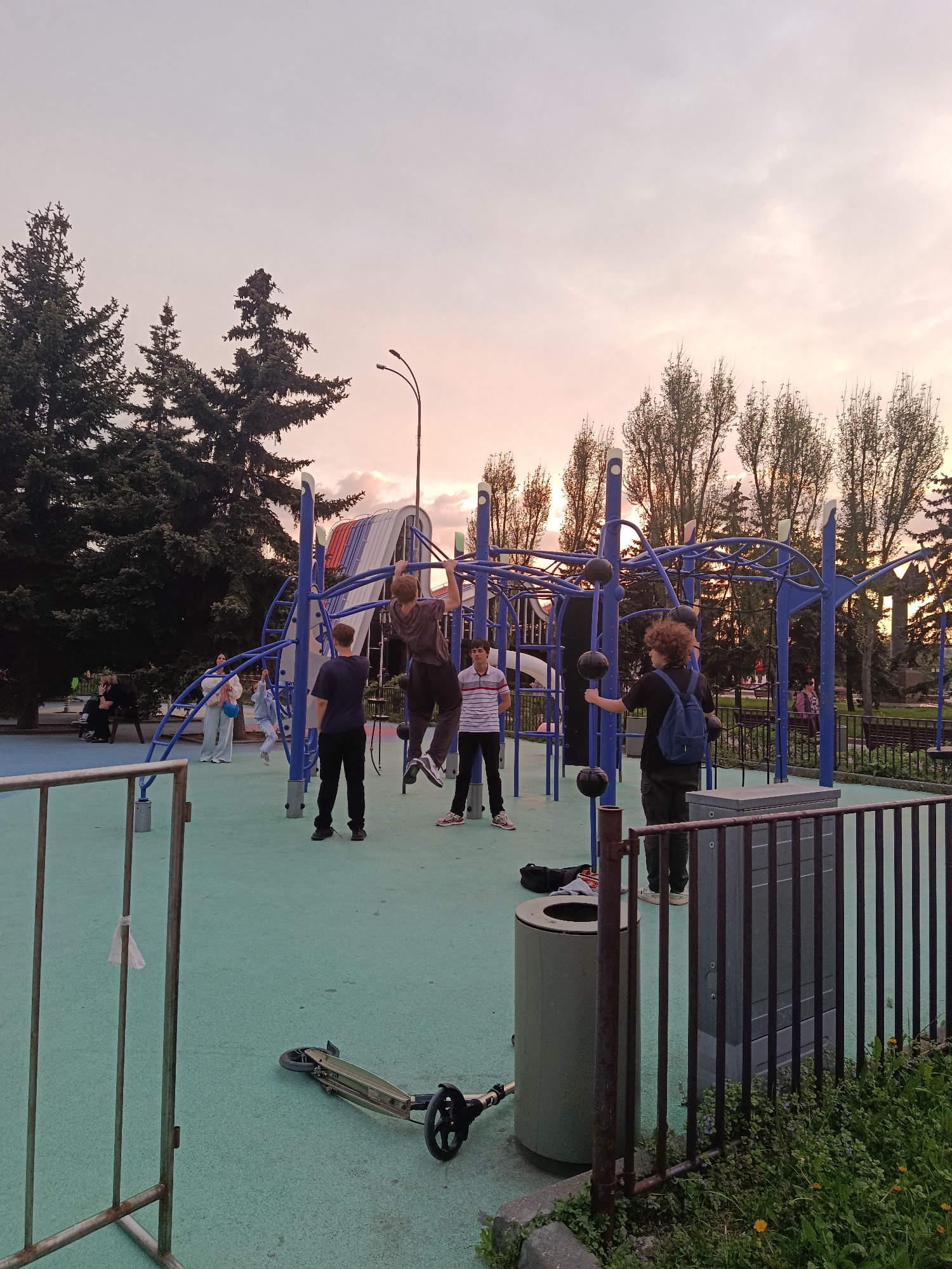The image shows a playground in Moscow, Russia. Several young people are present, with one climbing on blue metal play equipment. A few others are standing nearby. There is a metal fence surrounding the playground area. In the background, tall trees and a light post are visible under a pink-tinged sky. A scooter lays on the ground near a trash can.