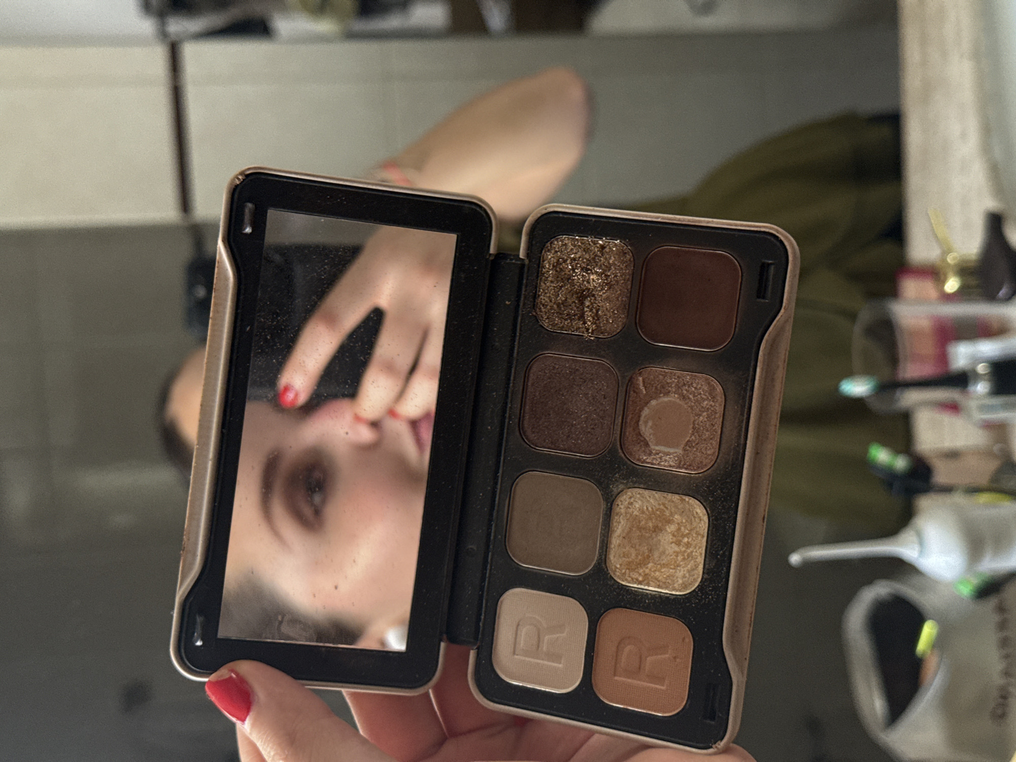 Here is a description of the image:
The image shows a person holding an open eyeshadow palette with a mirror on one side. The palette contains eight shades of eyeshadow in varying browns, golds, and neutrals. The mirror reflects the person's face. The person has red nail polish on their nails. The background is blurry but appears to be a bathroom setting. The specific location in San Pedro Garza García, Mexico cannot be determined from the image.