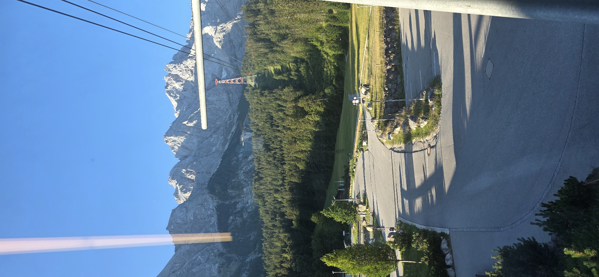 This is a vertical, eye-level, medium shot photograph taken from a moving cable car. The scene depicts a mountain landscape in Grainau, Germany, with a focus on the Zugspitze cable car infrastructure. In the foreground, a grey asphalt road curves through a parking area with several trees and large rocks. A tall, slender tree with dark green foliage stands on the left side of the road, casting a shadow. A concrete wall covered with grass and rocks lines the right side of the road.
In the midground, a dense forest of dark green pine trees covers a sloping hillside. A prominent red and white pylon, part of the Zugspitze cable car system, stands in the middle of the forest. The cable car's thick, grey support beam is visible in the upper midground, and three black cables extend diagonally across the sky.
The background features a majestic mountain range with jagged, rocky peaks, illuminated by the sun. The sky above is a clear, bright blue. The overall impression is a scenic view of the Bavarian Alps, likely taken during the daytime with sunny weather. There are no people or animals visible in the frame. The image is characterized by strong shadows, indicating a low sun angle, likely early morning or late afternoon.