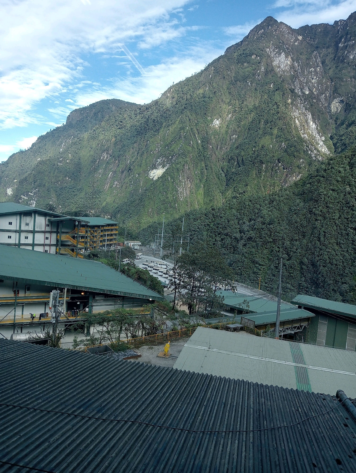 An aerial view captures a sprawling industrial facility nestled at the base of a massive, verdant mountain range in an undetermined region of Indonesia. The foreground is dominated by the corrugated metal roof of a large building, its dark gray surface weathered and lined. Beyond this, a cluster of industrial structures unfolds. Several buildings with green metal roofs and white facades, some featuring multiple stories with exterior staircases painted bright yellow, are arranged in a complex. Adjacent to these buildings, a vast parking lot is visible, filled with numerous white buses and smaller vehicles. Power lines and utility poles are scattered throughout the scene, indicating the operational nature of the complex. The mountain behind the facility is steep and covered in dense, dark green foliage, with rocky outcrops and streaks of lighter-colored mineral deposits visible on its surface. The sky above is a mix of blue and white clouds, with faint contrails suggesting aircraft activity. The overall impression is one of an industrial outpost operating within a rugged, natural environment.