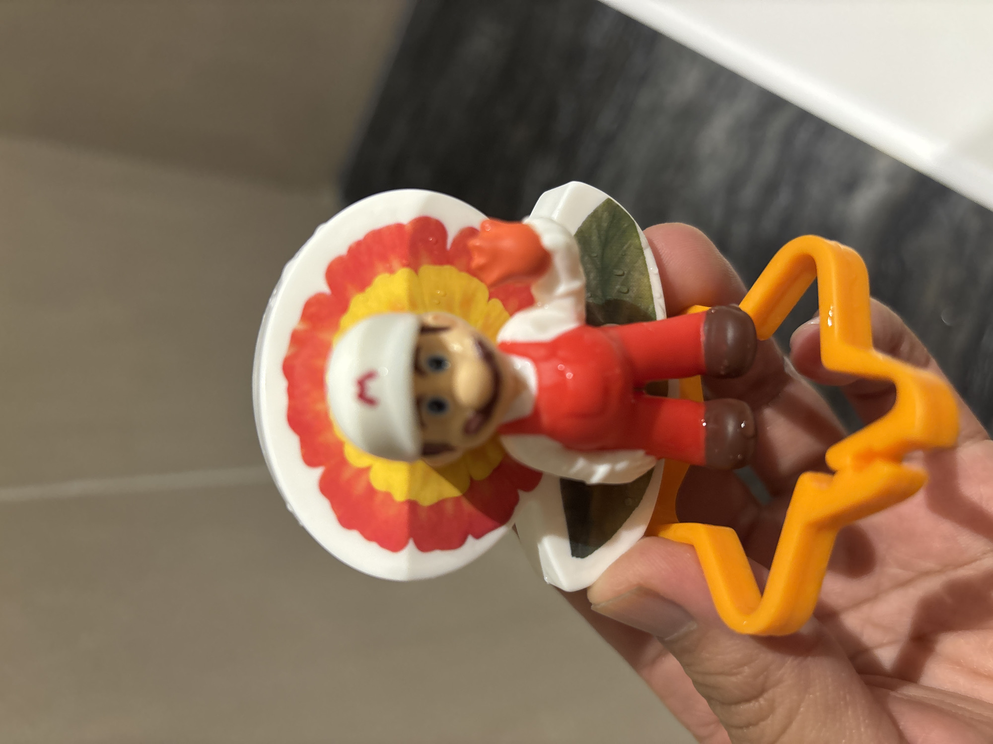 A close-up shot shows a hand holding a Mario figurine, part of a toy. The figurine is wearing red overalls and a white hat with an 'M' logo. Behind the figurine, a round, white object is decorated with a colorful, petal-like design in red, yellow, and orange. The base of the toy is an orange, star-shaped plastic piece. The background is out of focus, showing a beige wall on the left and a dark, textured surface, possibly a floor, on the right. A bright white surface is visible on the far right. The lighting appears to be artificial and bright. The scene is indoor, with no indication of location in Dubai, United Arab Emirates.