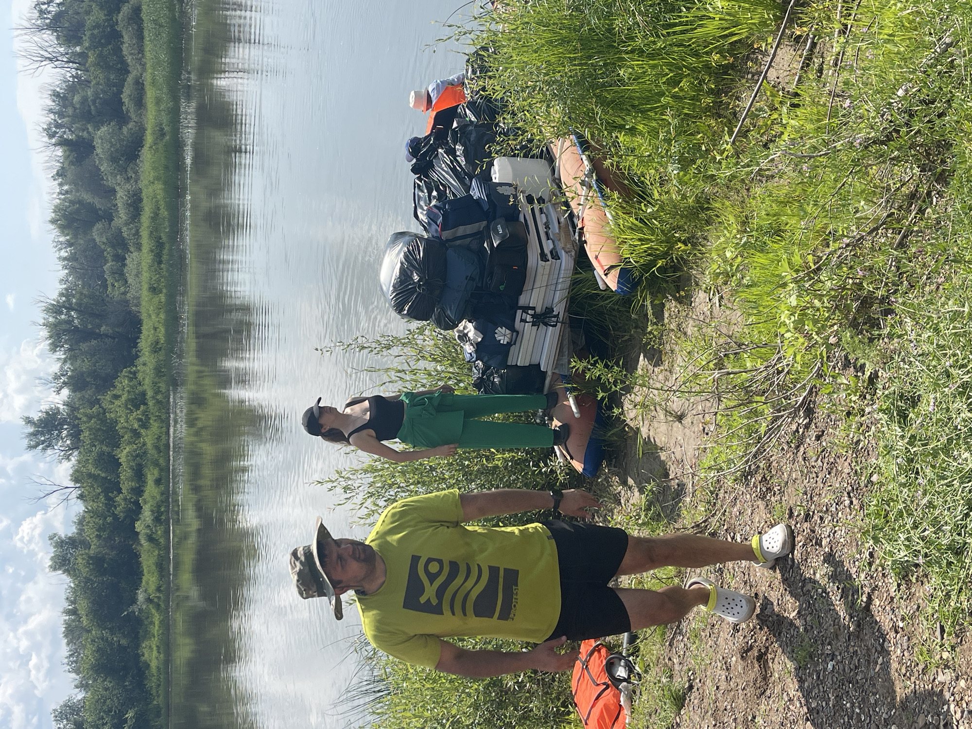 This image captures a scene by the river in the city of Nezhinka, Russia. In the foreground, a man wearing a camouflage hat, a lime green t-shirt with a graphic design, and black shorts is walking towards the viewer. He is also wearing white Crocs. To his left, on the riverbank, is an orange life vest. Behind the man, a woman stands near a large pile of gear on a small cart. She is wearing a black tank top and green pants, with a black baseball cap. The gear pile consists of several large black trash bags, duffel bags, a white plastic container, and what appear to be folded white tables. An inflatable raft lies on the ground in front of the cart. The background shows a wide, calm river reflecting a cloudy sky and a dense line of trees and vegetation along the opposite bank. The overall environment is natural and appears to be a staging area for an outdoor activity, likely involving the river.