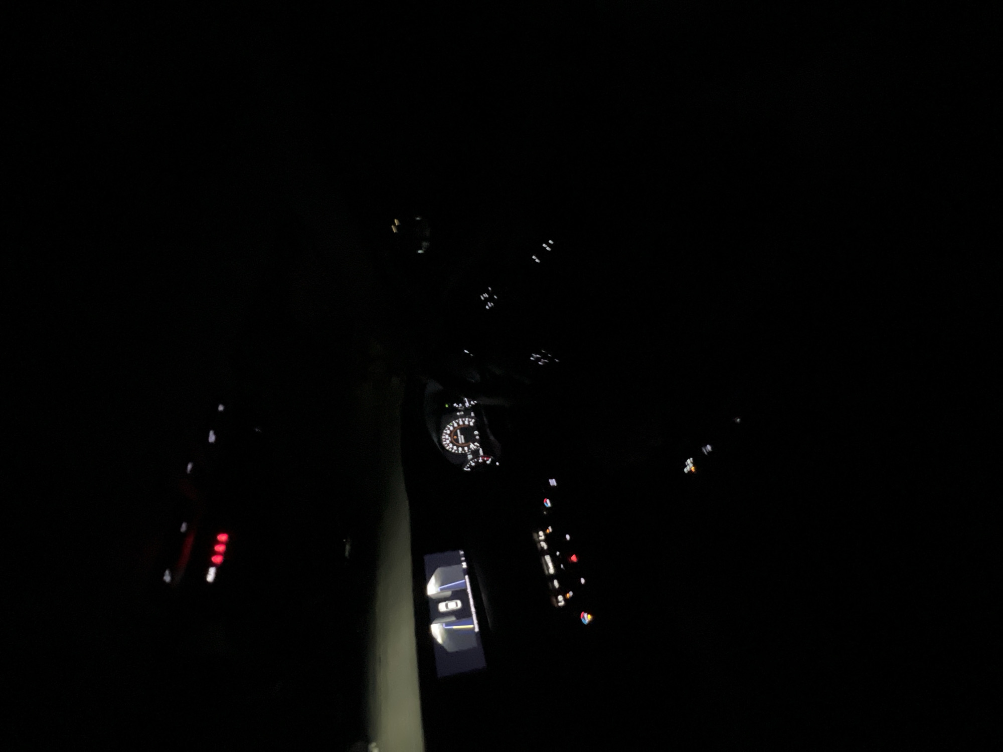 The image captures the interior of a car at night, viewed from an elevated angle, likely from the passenger side or rear. The scene is dominated by darkness, with various illuminated elements of the vehicle's dashboard and center console providing the only light sources.
The central focus is on the instrument cluster, visible in the upper-middle section, displaying a circular gauge with white backlighting, resembling a speedometer or tachometer. Below and slightly to the left of the instrument cluster, a digital screen on the center console is illuminated, showing a graphic representation of a white car from a top-down perspective, surrounded by lines, strongly suggesting a parking assist system or a 360-degree camera view is active.
To the left of the center console, a cluster of red and white illuminated buttons or indicators is partially visible. On the right side of the center console, numerous other smaller buttons and indicators are lit up in various colors, including white, orange/yellow, and red, hinting at controls for climate, media, or other car functions. The overall appearance suggests a modern car interior.
The time of day is clearly night, as indicated by the complete darkness surrounding the illuminated controls. No individuals are visible in the image. No discernible text is clearly legible on any of the displays or buttons due to the angle and resolution. The activity taking place is inside a powered-on vehicle, possibly stationary or in motion.