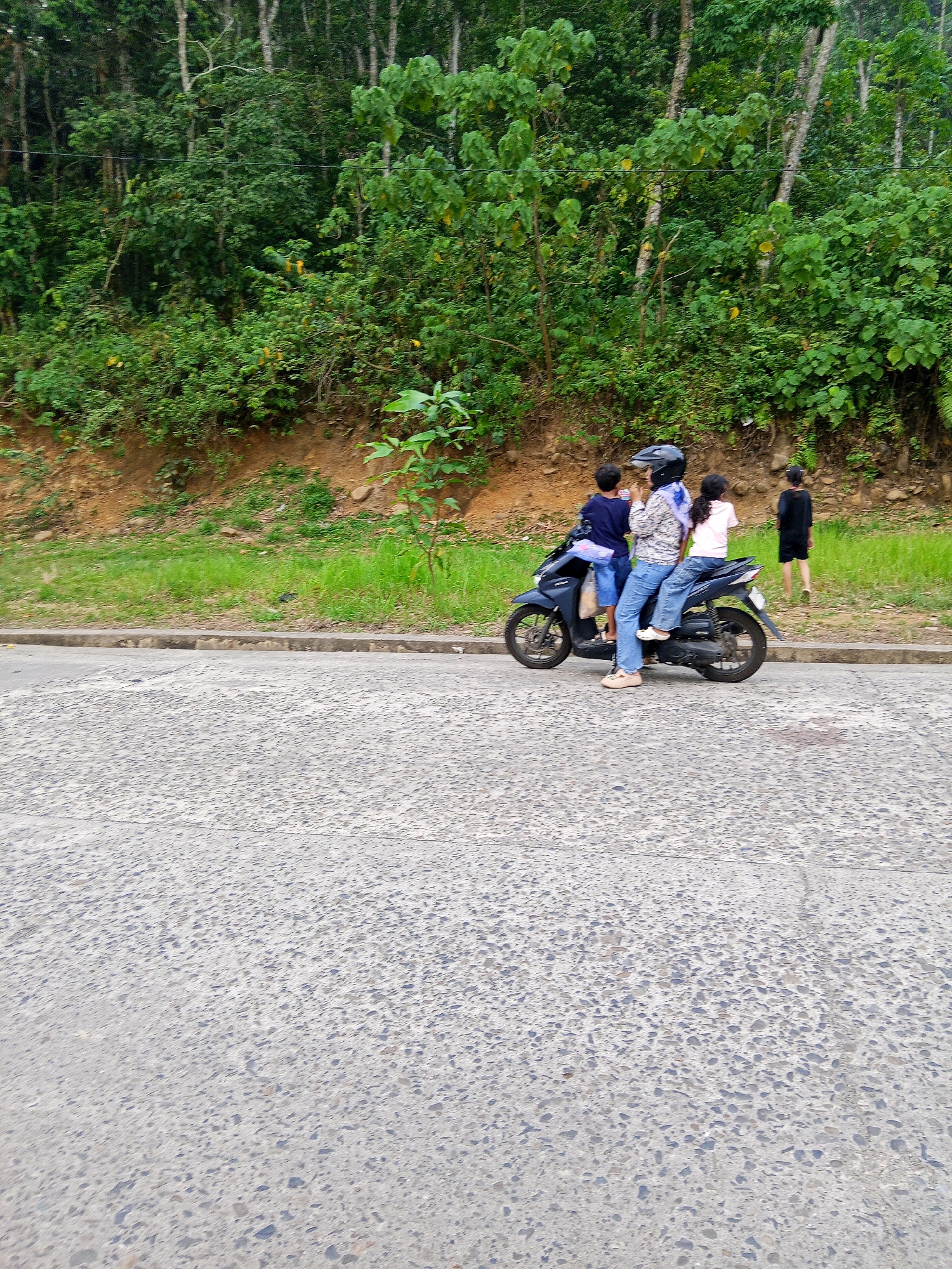 In Lubuklinggau, Indonesia, a family is seen riding a motorcycle. A woman wearing a helmet and a patterned shirt, with a blue scarf, is driving the motorcycle. She is accompanied by two children, a boy in a blue shirt and blue shorts, and a girl in a pink shirt and blue shorts, both sitting behind her. Another child, dressed in black, stands on the right side of the image, observing. The family is on a road bordered by lush greenery, with a dirt embankment and dense trees in the background. The road surface is asphalt with a textured finish.