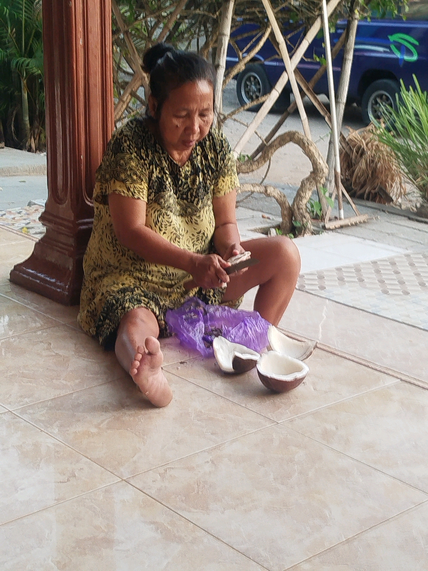 A woman is sitting on a tiled patio in Burneh, Indonesia, preparing to open coconuts. She is wearing a yellow and black patterned dress and has her hair pulled back in a bun. She is holding a knife in her right hand and appears to be in the process of cutting into a coconut. Several opened coconut halves and a purple plastic bag are on the ground next to her. In the background, a blue van is visible, along with some bamboo structures and vegetation. The scene suggests a domestic setting where food preparation is taking place outdoors.