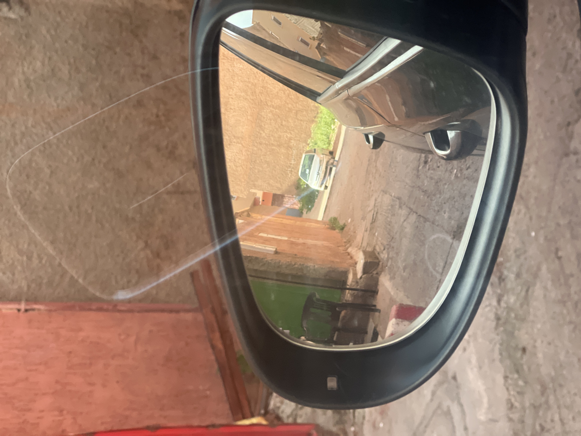 The image shows the reflection in a car's side mirror. The mirror reflects a narrow street in Ouaouizert, Morocco, lined with brick buildings and stone pathways. A white car is partially visible next to the reflecting car. In the background, more buildings and vegetation are visible. The image appears to be taken from inside the car. A red object is visible near the bottom left of the mirror's frame. A thin, translucent line crosses the view in the area around the left of the mirror, it could be a scratch on the window. A small white reflection is also visible on the mirror's surface.