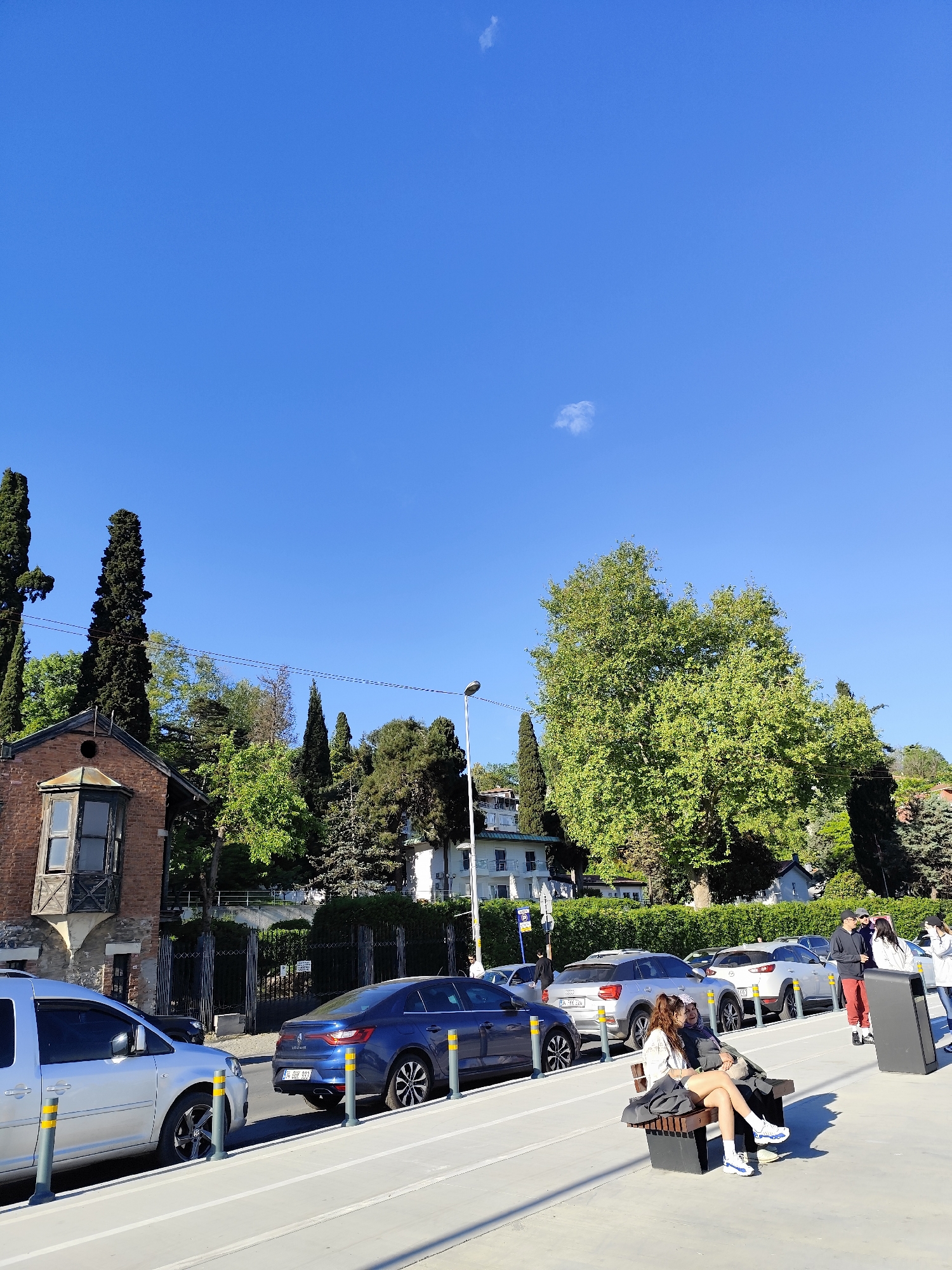 The image captures a street scene in Sarıyer, Turkey on a sunny day with a clear blue sky. Along the road, cars are parked, including a silver van and a blue sedan with the license plate "34 SHH 593". A gray SUV and a white car are also visible further down the street.
A wooden bench sits beside the road, where two people are seated. Nearby, a group of people are standing and talking. A trash receptacle is positioned near the group.
The background features a mix of architecture and nature. A brick building with a distinctive window is on the left, followed by a variety of trees, including tall evergreens. A white building is nestled among the trees. A street lamp is also visible in the background. The setting conveys a blend of urban and natural elements in Sarıyer.