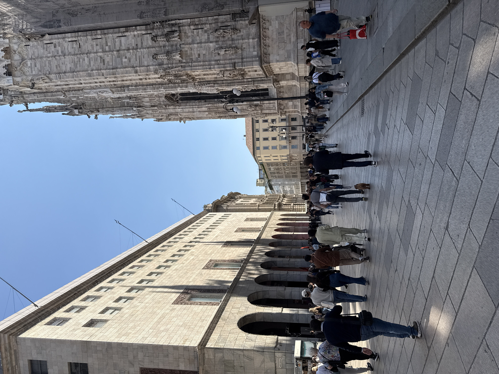The image shows a crowded street scene in Milan, Italy, likely near the Duomo di Milano (Milan Cathedral). A long line of people stretches along the paved walkway between the cathedral and a large, light-colored building with arched colonnades. The sky is clear and blue. The people are mostly dressed in casual clothing, with many wearing backpacks or carrying bags. The architectural details of the cathedral are visible, including its spires and intricate stonework. The building opposite the cathedral features rows of windows and arched openings. The overall impression is of a busy tourist area.