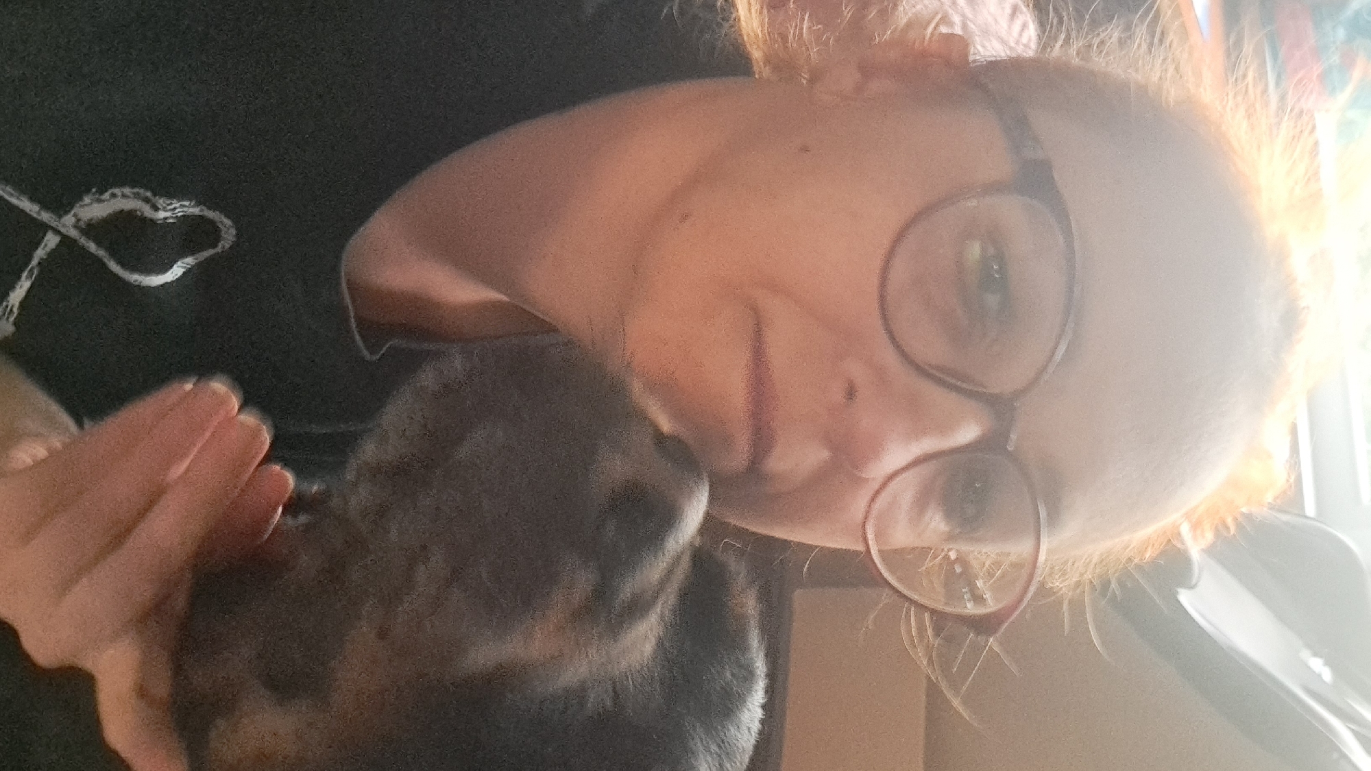 The image shows a close-up, selfie-style view of a person with reddish-blonde hair, wearing glasses and a dark t-shirt with a white ribbon design. The person's face is partially obscured by the head of a dog, which is nuzzling against their cheek. The lighting suggests an indoor environment, possibly a home, with a window or bright light source in the background. The overall impression is one of affection between the person and the dog. There is no discernible information within the image that indicates a specific location in the city of Riga, Latvia.