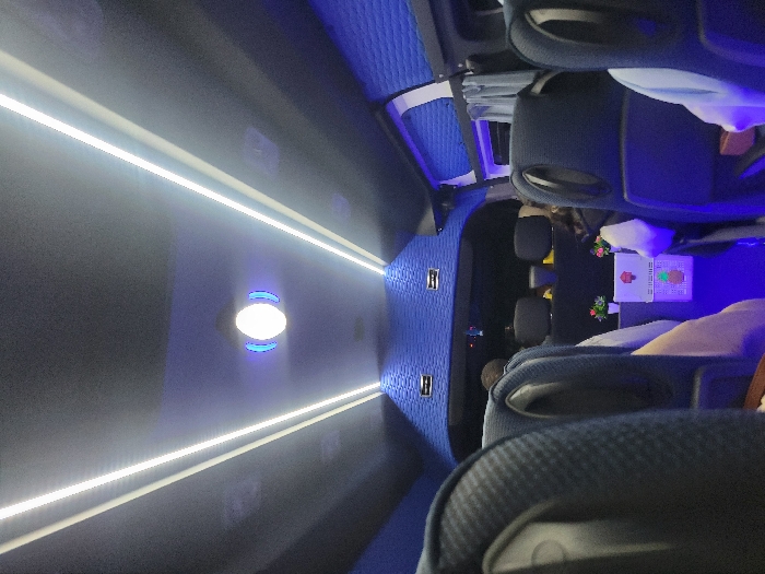 An interior shot of a passenger vehicle like a bus or limousine. Rows of dark-colored, plush seating with headrests are visible. Overhead, long, parallel light strips illuminate the space, and circular lights are mounted in the center. The ceiling and walls are blue with a textured pattern. A few passengers are seated in the rows.