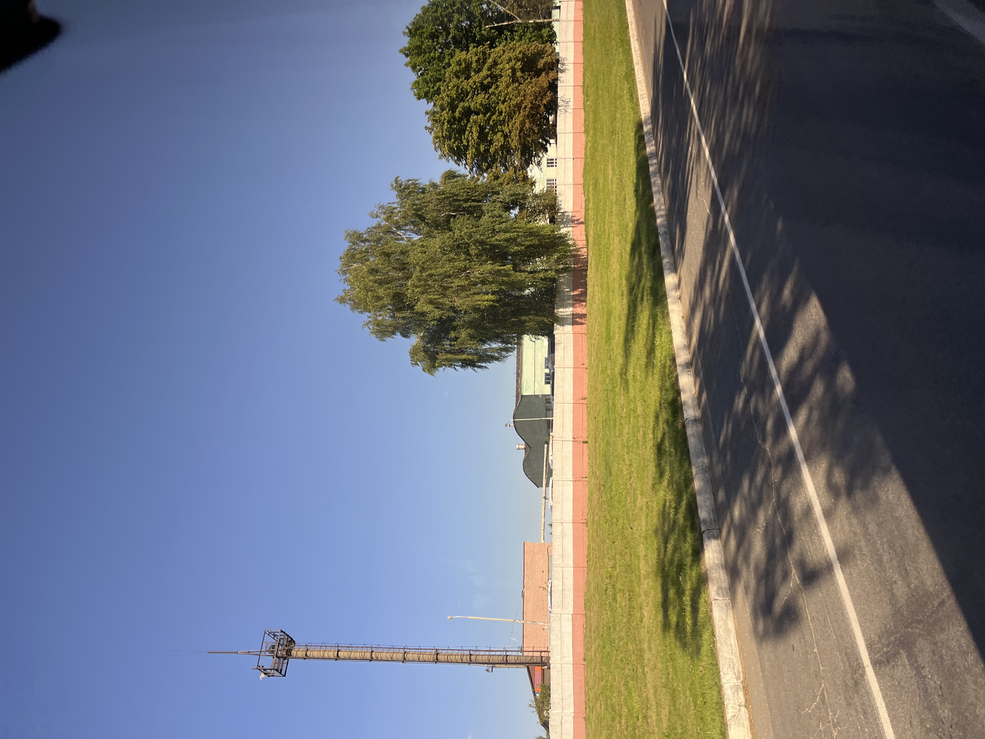 The image depicts a roadside view in the city of Myadzyel, Belarus, on a clear day with a bright blue sky. In the foreground, a dark asphalt road with a white line is visible, casting dappled shadows from unseen trees. To the left of the road is a strip of green grass bordered by a concrete curb. Beyond the grass, a long, low concrete fence with a red and white striped pattern separates the road area from a grassy field.
Further back, various industrial-looking buildings are visible. To the far left stands a tall, cylindrical industrial smokestack with a metal platform at its peak. Next to the smokestack is a low, rectangular brick building. In the center background, there are two multi-story buildings with light-colored facades and dark roofs. To the right of these buildings, a cluster of deciduous trees with green and some yellowing foliage stands. The overall scene suggests an industrial or commercial area situated on the outskirts of the city. There are no people or animals visible in the image.