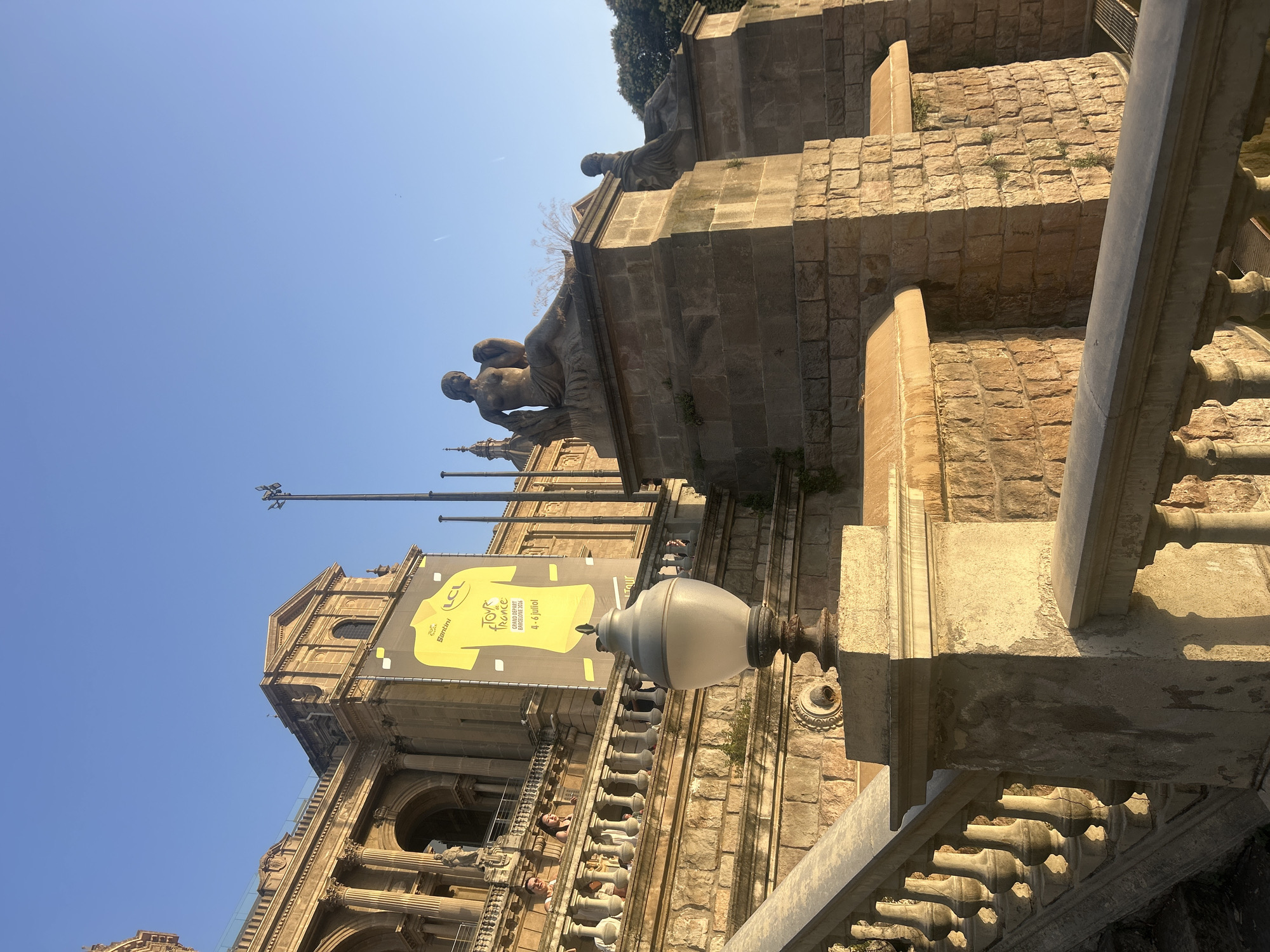 The image depicts a section of the Palau Nacional, a monumental building located on Montjuïc hill in Barcelona, Spain, which houses the National Museum of Catalan Art. The perspective is from a grand stone staircase leading up to the Palau. In the foreground, a stone balustrade and part of a lamp post are visible. The middle ground shows more of the ornate stonework of the staircase and terraces, with several stone statues perched atop prominent plinths. Behind these statues, a large banner featuring a yellow cycling jersey with the text "Tour de France" and "Grand Départ Barcelona" is prominently displayed against the facade of the Palau Nacional. The building itself exhibits neoclassical architecture with grand columns and arched entrances. The sky above is a clear, bright blue, indicating a sunny day. People are visible on the terraces and the staircase, suggesting it is a public space actively being visited. The lighting suggests it is either early morning or late afternoon, with the sun casting long shadows.