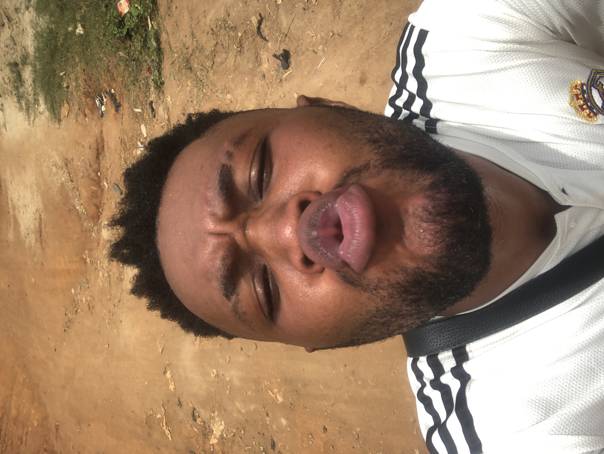 A man with dark curly hair and a beard is making a pouting face with his tongue slightly sticking out. He is wearing a white sports jersey with black stripes on the shoulder and a black strap across his body. The background consists of dirt and some sparse vegetation. The location is Unknown, Unknown.