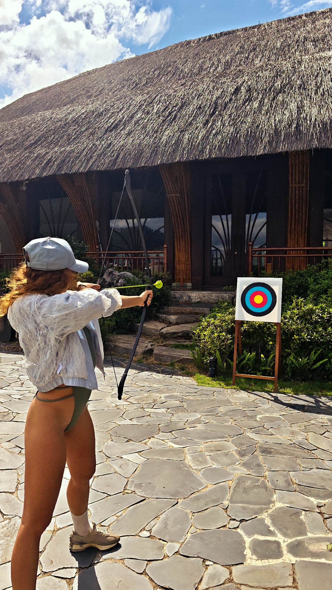 An outdoor, sunlit photograph captures a woman engaged in archery, aiming at a target in Púng Luông, Vietnam. The woman is viewed from her back-right side, wearing a light-colored cap, a textured light jacket or shirt, green bikini bottoms, and sneakers. Her curly red hair is visible beneath her cap. She holds a black bow, drawing back a yellow, possibly foam-tipped, arrow, poised to shoot.
In front of her, to the right of the frame, stands a white archery target board with concentric circles of red, yellow, blue, black, and white, mounted on a simple wooden stand. The ground beneath her and extending throughout the foreground is paved with large, irregularly shaped flagstones.
The background features a large, traditional-style building with a distinctive, thick thatched roof. The building's structure includes dark wooden or bamboo-like pillars and window frames. Steps crafted from natural stone lead up to the building's entrance. Lush green bushes and foliage are visible around the building and the target area. The sky above is bright blue with scattered white clouds, indicating clear, sunny weather, likely midday or early afternoon. The scene conveys an atmosphere of leisure and recreational activity in a tropical or resort-like setting. There is no visible text.