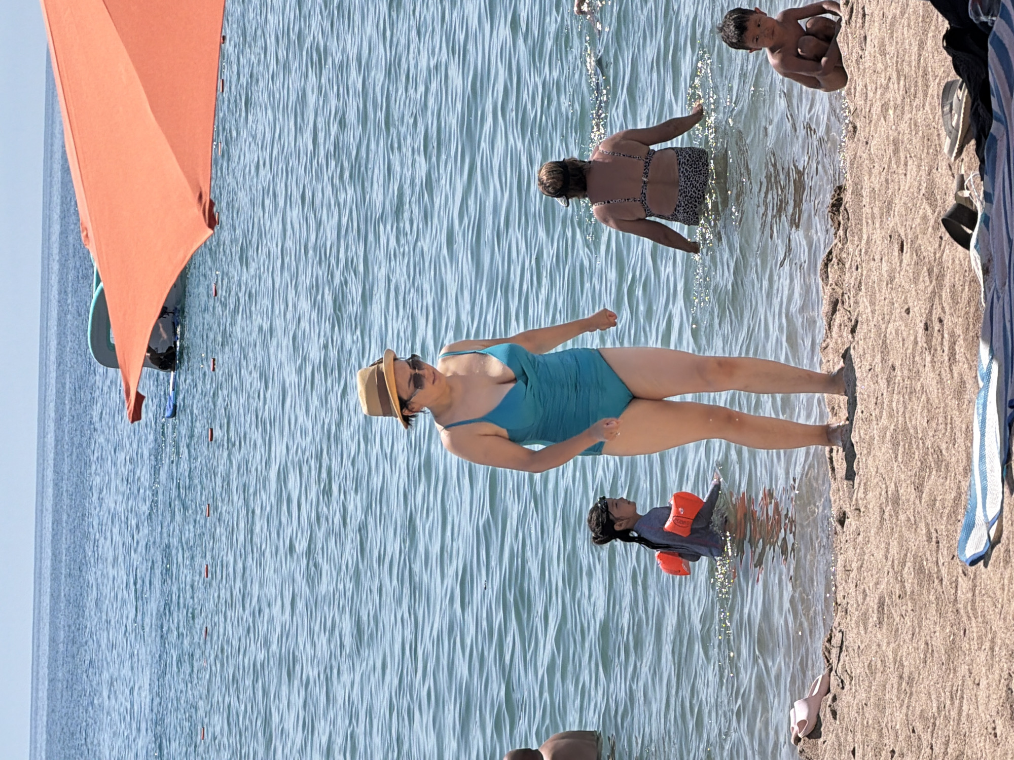 This is a photograph of a beach scene on a sunny day. The image features several people enjoying the water and sand. In the foreground, a woman in a turquoise swimsuit and a straw hat stands in the shallow water, facing away from the camera. To her left, a child in a grey long-sleeved shirt and orange floaties is also in the water, looking towards the woman. In the background, another woman in a patterned bikini is wading in the water. Further back, a young boy is crouched on the sandy shore.
The water is clear with gentle waves, reflecting the sunlight. The beach is sandy, and a portion of an orange beach umbrella is visible on the left side of the frame. There are also some beach towels and clothing items on the sand. The overall atmosphere appears relaxed and leisurely, suggesting a typical summer day at the beach. The time of day is likely daytime, given the bright sunlight and the shadows. The location is not specified but appears to be a coastal area. No visible text is present in the image.
