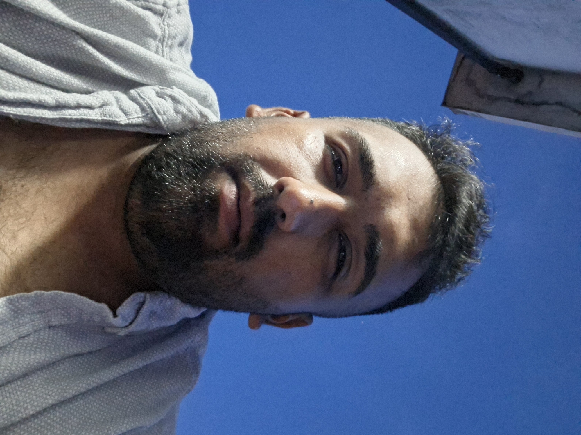 The image is a close-up, low-angle selfie of a man against a bright blue sky. The man has dark, curly hair, a beard, and is wearing a light-colored, textured shirt. A portion of a grey, textured structure is visible in the upper left corner of the image, possibly a building or overhang. The man's face is turned slightly towards the camera, and he appears to be outdoors in an urban setting, potentially Evere, Belgium, given the blue sky and architectural element.