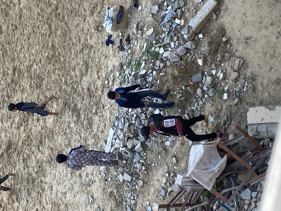 The image depicts an outdoor scene in Khan Yunis, Palestinian Territory, characterized by a mix of sandy terrain and rubble. Four males, appearing to be children or adolescents, are present.
The upper portion of the scene is dominated by light brown, undisturbed sand. A male with only his lower legs visible is walking left in the upper left corner. In the upper center, another male, barefoot, wearing a dark hooded top and shorts, walks right.
The lower two-thirds of the image consists of a rough, uneven ground covered extensively with construction debris, including concrete blocks, broken bricks, and scattered smaller stones. Small patches of green vegetation are interspersed among the rubble.
Standing in the mid-left area, facing away from the viewer towards the sandy expanse, is a male wearing a patterned grey and black tracksuit with brown shoes. To his right, in the mid-right, a male wearing a dark blue long-sleeved shirt with lighter blue accents, blue jeans, and dark athletic shoes walks left.
In the lower center-left, another male wearing a black cap, a black vest over a maroon long-sleeved shirt (with "FOREVER BALENCE" visible on the left sleeve), black trousers, and reddish-brown slip-on shoes walks left, holding a small dark object in his right hand. Adjacent to him, in the lower left, is a collapsed or dismantled structure of metal beams, partially covered with a white canvas sheet.
On the far right, amidst the rubble, are several pairs of sandals and shoes (blue and brown) along with some folded clothing (a blue shirt and white fabric) placed on or next to a broken concrete block.