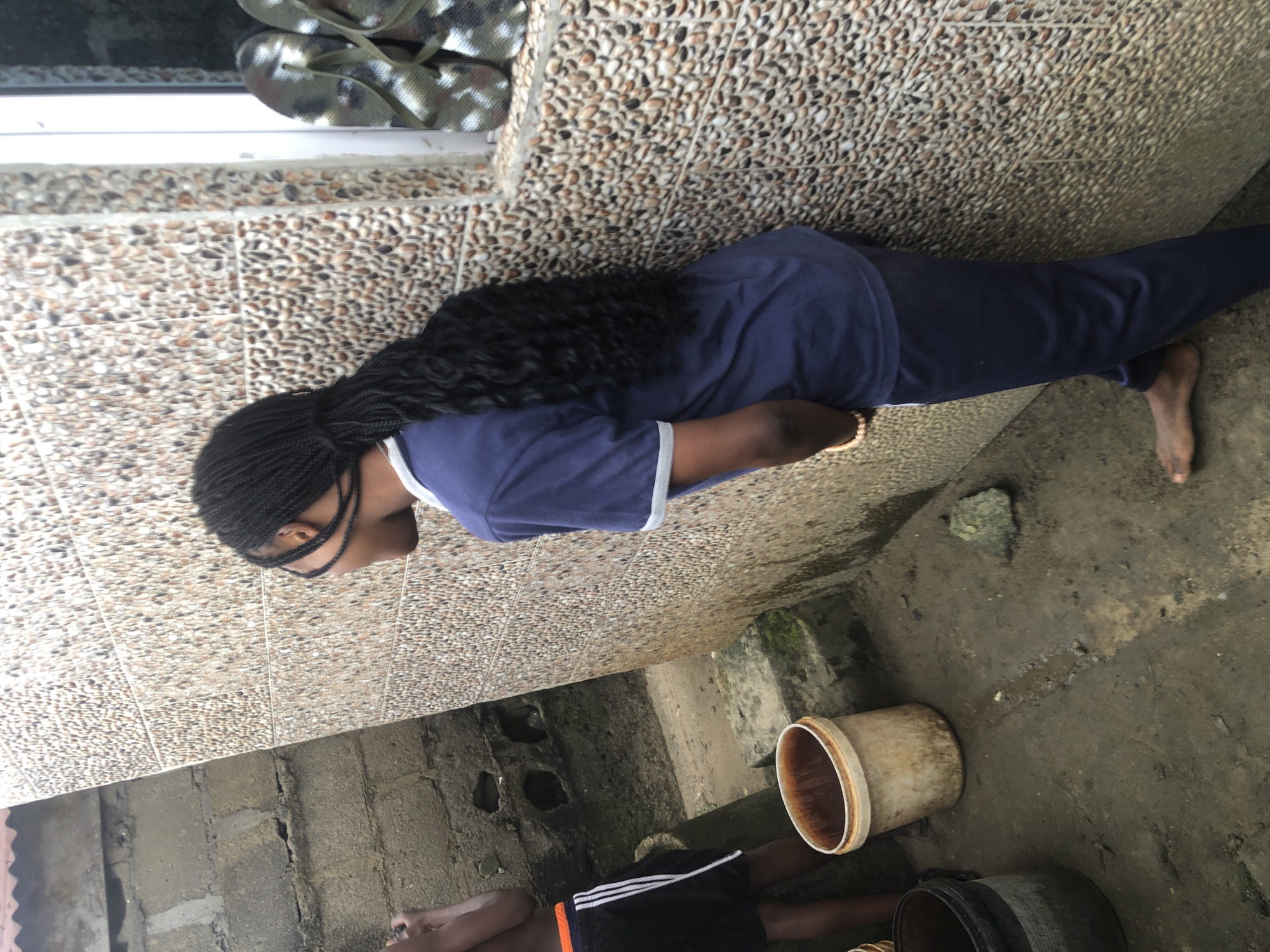 The image shows a young Black person with braided hair, wearing a navy blue tracksuit, standing with their back to the camera against a pebble-tiled wall. They are in an outdoor environment with a rough, wet ground surface. To the left of the frame, another Black person is partially visible, wearing black shorts with white stripes and an orange waistband. Two buckets are present on the ground, one beige and stained, the other dark grey and black. In the background, a rough concrete block wall is visible. On the ledge of the pebble-tiled wall, a pair of camouflage flip-flops are placed. The scene appears to be in a less developed urban area, possibly Ado, Nigeria, given the context.