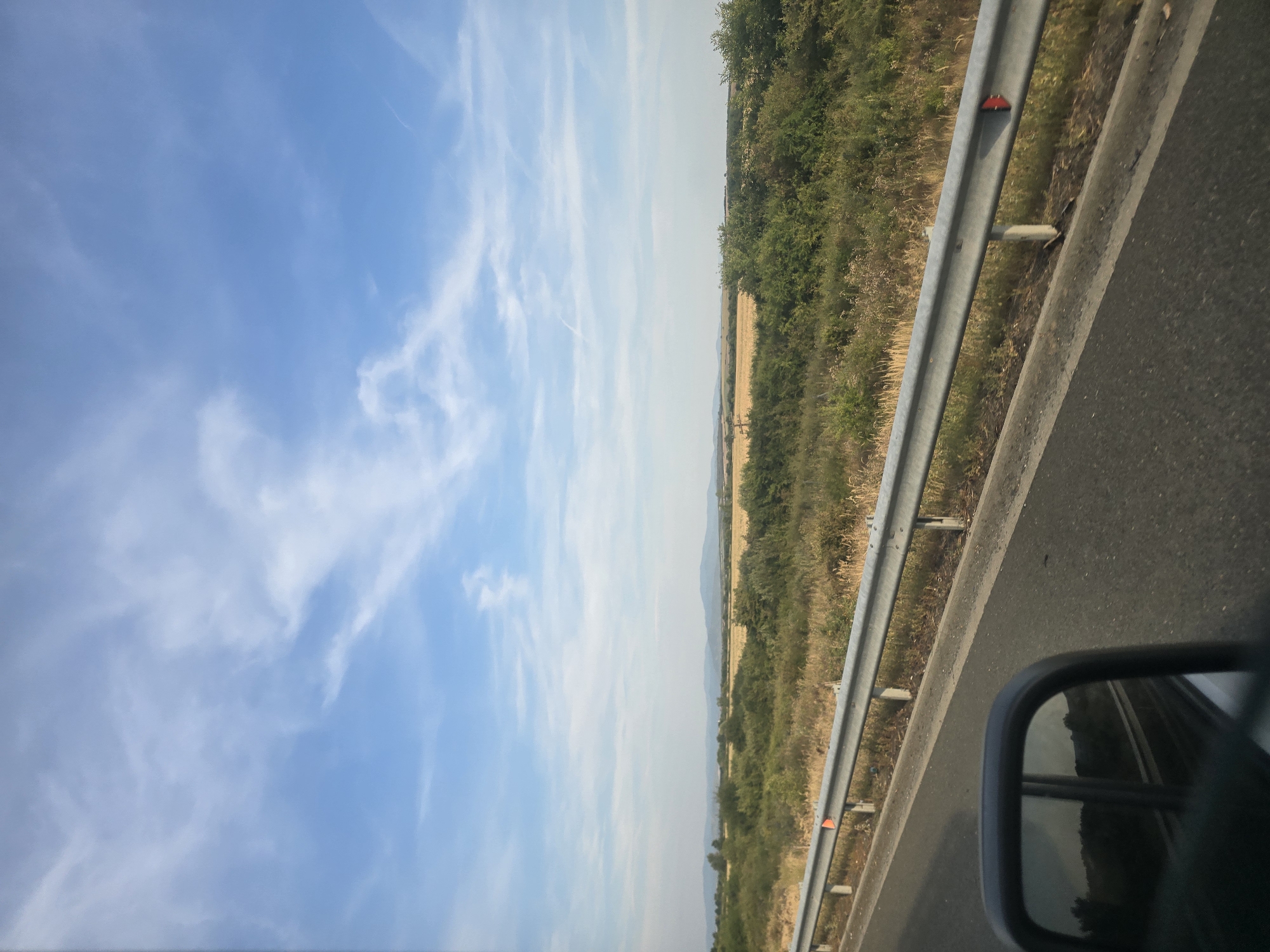 The image shows a view from a moving vehicle, likely a car, traveling on a road with a metal guardrail on the right side. The road surface is asphalt. Beyond the guardrail, there is a wide expanse of dry grass and low-lying green vegetation. Further in the distance, there are fields of golden crops, possibly wheat or barley, and then rolling hills or mountains under a bright blue sky with scattered white clouds. The scene appears to be in a rural or semi-rural area. There are no people or animals visible in the frame. The reflection in the car's side mirror shows a similar landscape, indicating the direction of travel. The image has been identified as being from Cutina, Romania.