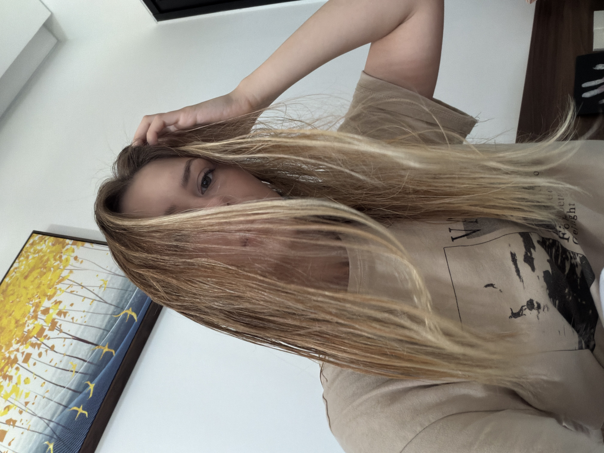 A young Caucasian female with long blonde hair is looking at the camera in a selfie-style shot. Her hair is partially covering her face, with strands falling over her eyes. She is wearing a light brown t-shirt with a graphic print of silhouetted figures on a landscape. Her right hand is raised, touching her hair near her temple. The background consists of a white wall, a framed painting with yellow and blue abstract elements, and a dark wooden headboard of a bed. The lighting suggests an indoor setting, possibly a hotel room, and the overall mood is casual. The image does not contain any information that would place it in Đà Nẵng, Vietnam.