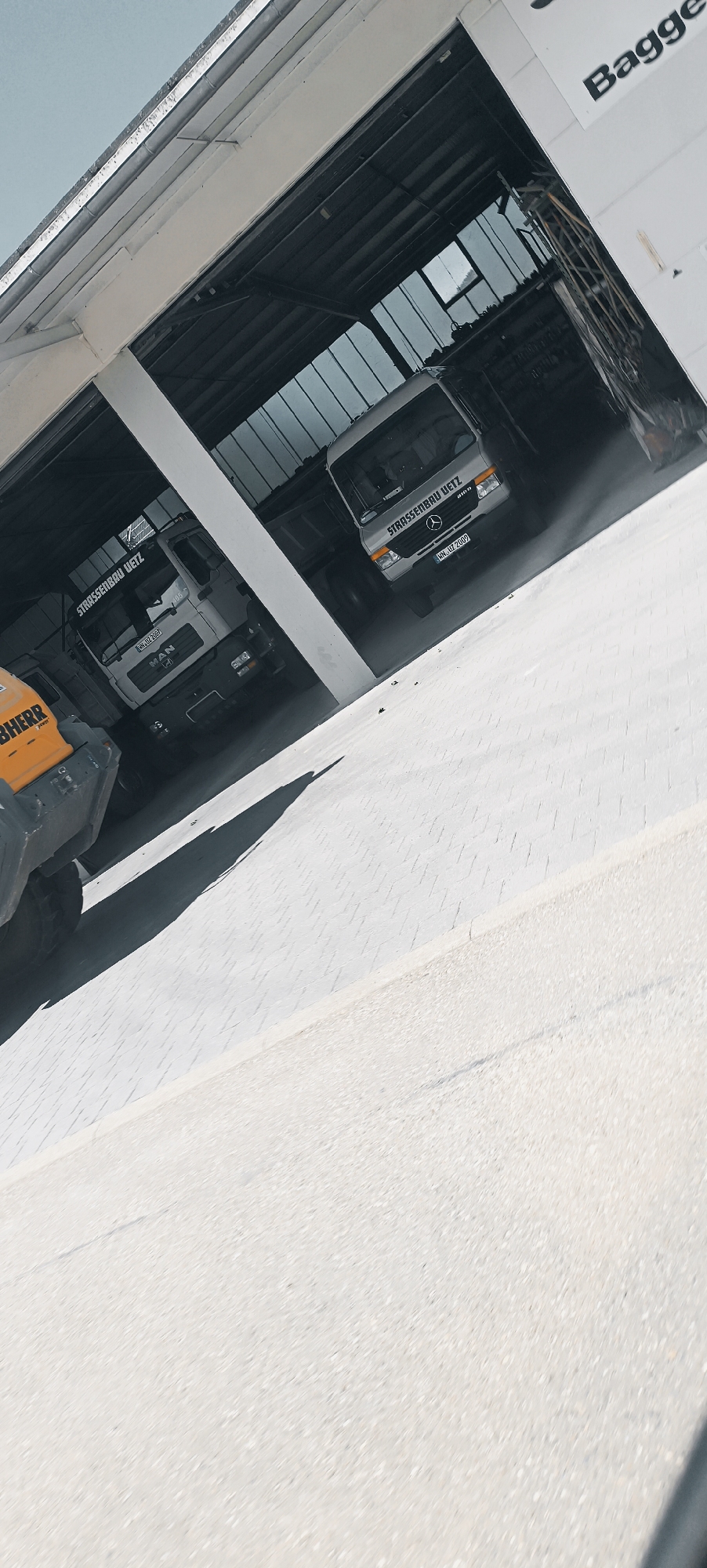 The image depicts a scene at a construction site, likely a depot or garage, in the city of Winterbach, Germany. The primary subjects are two white trucks and a yellow construction vehicle. The truck on the left has "MAN" branding on its front. Both trucks have "STRASSENBAU UETZ" written on their sides, indicating they are part of a road construction company. The truck on the right also has a Mercedes-Benz logo. The yellow vehicle appears to be some form of heavy machinery, possibly a loader, with "LIEBHERR" branding visible.
The trucks are positioned inside a large, open-sided building with a metal roof supported by white beams. The building's floor is paved with light-colored interlocking blocks, and a similar surface extends outside. The building has large windows or openings, allowing natural light to illuminate the scene and offering a view of the sky. The sky itself is a pale blue.
The perspective is from a low angle, looking up towards the building and the vehicles. The overall impression is one of industrial activity, with the vehicles presumably parked or undergoing maintenance. There are no people clearly visible in the image. The environment suggests a typical day at a construction company's facility, with the vehicles and building being the main focal points.