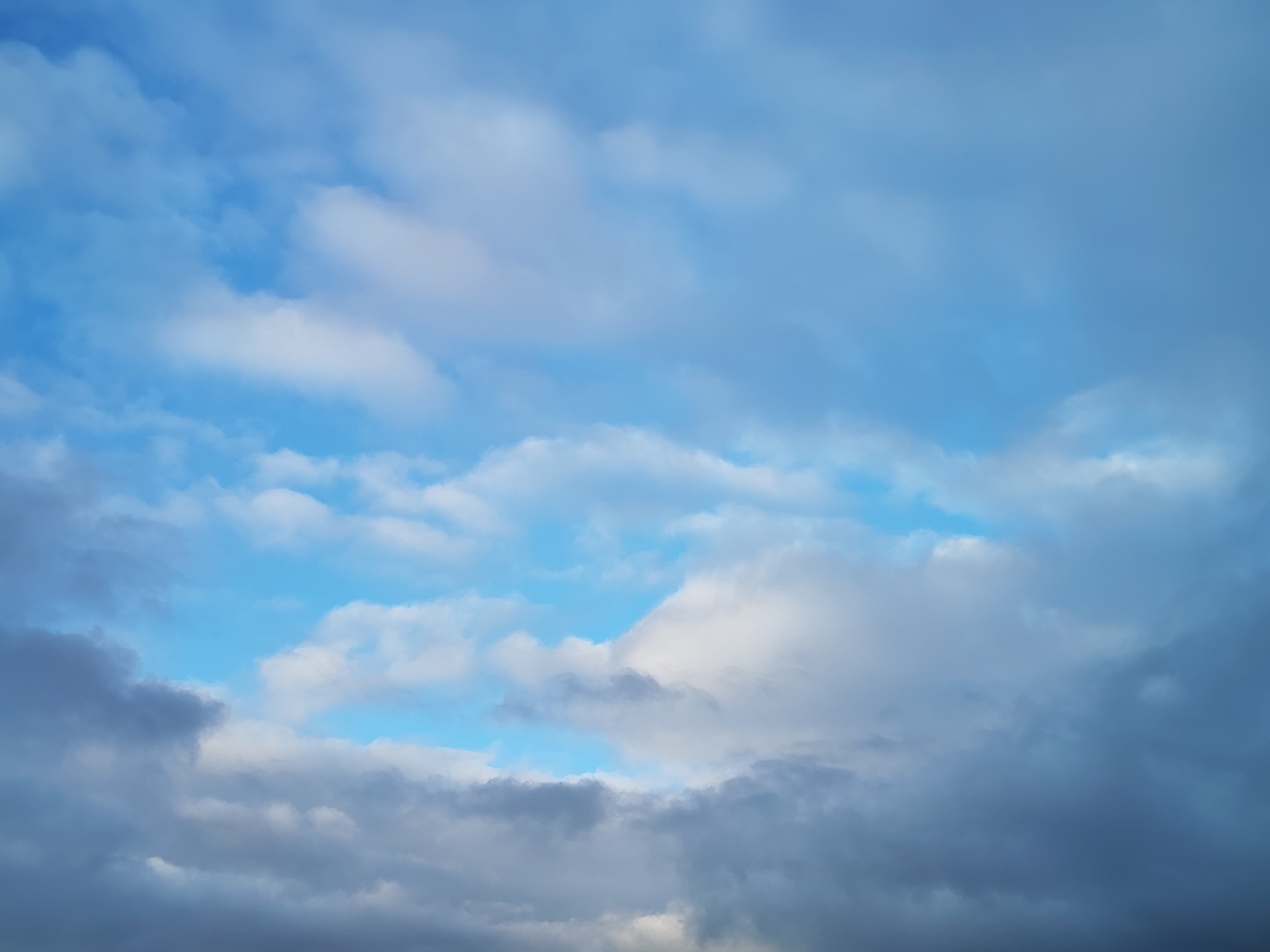The image captures a dynamic sky scene, primarily featuring clouds against a blue background. The sky displays a varied palette, ranging from vibrant, clear blue in areas where the sun is breaking through, to lighter blues, and progressively darker shades of grey and bluish-grey where clouds are denser.
The subjects are exclusively atmospheric elements: an expansive sky and numerous clouds. There are no people, objects, or structures visible.
The activity depicted is the natural formation and presence of clouds in the sky. Some clouds appear bright white and fluffy, suggestive of cumulus formations, particularly in the upper and central parts of the image where the blue sky is more prominent. In contrast, the lower and right sections feature heavier, darker, and more uniform cloud masses, resembling stratocumulus or nimbostratus clouds, which often indicate an overcast condition or approaching precipitation.
Notable details include the time of day, which appears to be daytime, likely a transitional period such as mid-morning or late afternoon, given the blend of bright light and shadowed cloud masses. The weather is characterized by a mix of partial clearing and heavy cloud cover, suggesting variability. No specific location cues are visible within the image itself that would identify Izhevsk, Russia. The scene evokes a sense of natural movement and atmospheric depth. There is no visible text in the image.