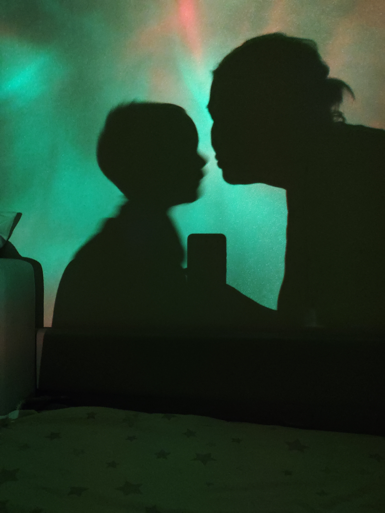 A shadow of two figures, likely a parent and child, are projected onto a surface with green and red lights in the background. The child is holding a phone in their hands. A section of a bed or sofa is visible below the shadows. There is no definitive information to locate the photo in Markovskii, Russia, the photo gives no evidence for location.
